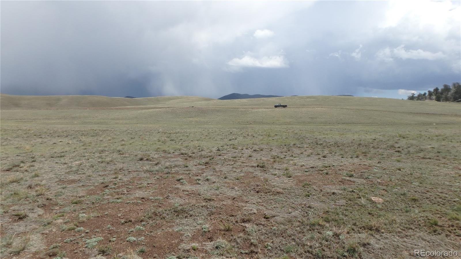 Open grassland with rolling hills, distant mountains, and a pickup truck beneath storm clouds.