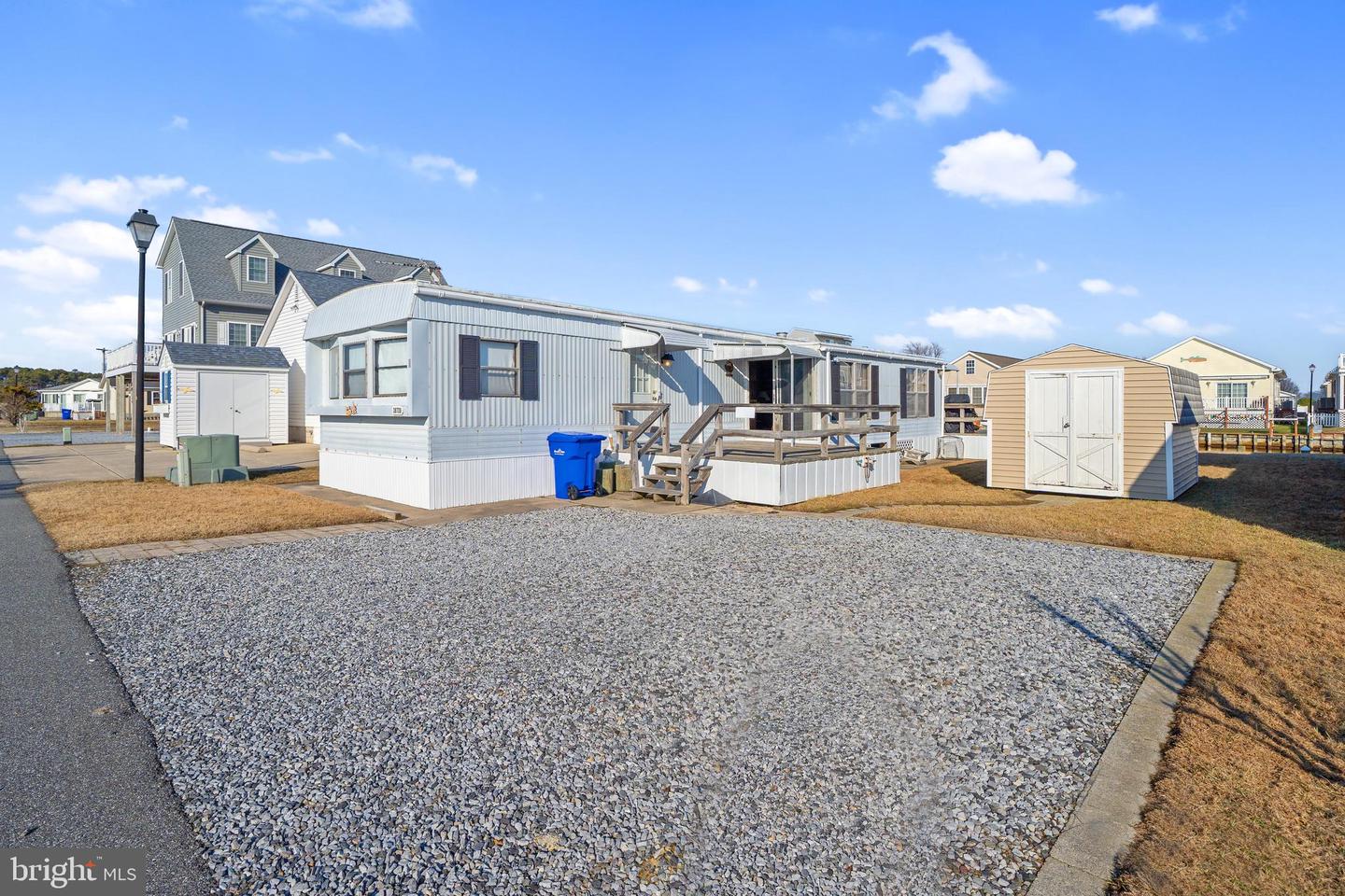 Manufactured home with a raised wooden deck beside a large gravel parking area and white storage shed.