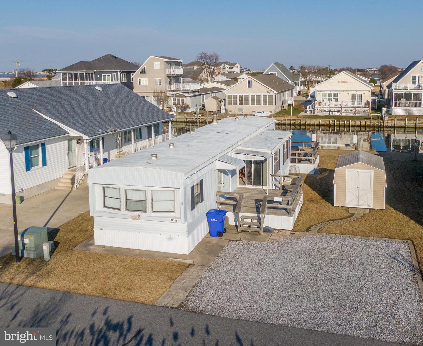 White manufactured home with a wooden deck and gravel driveway beside a canal.