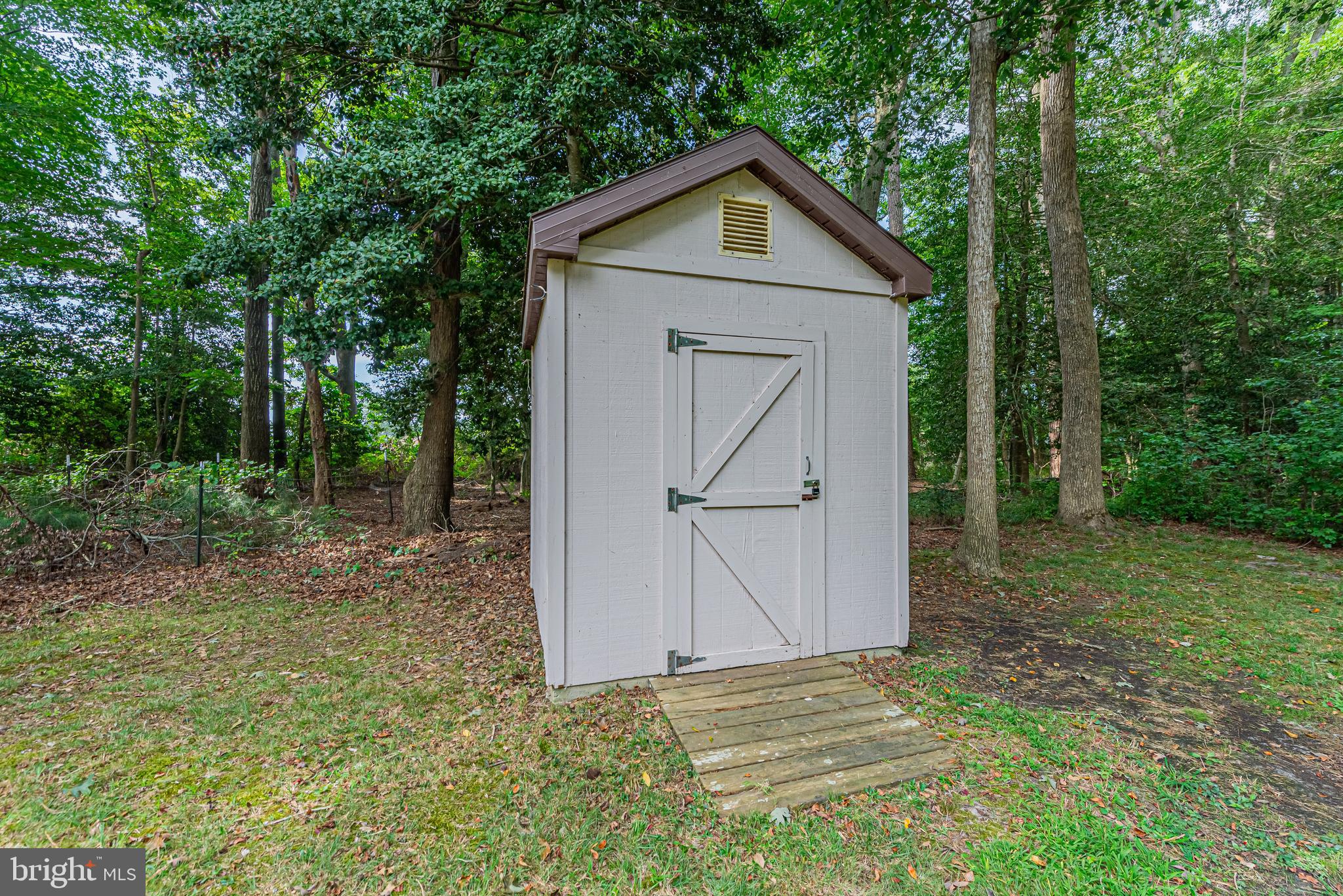 White storage shed with brown trim and a wooden walkway in a wooded yard.