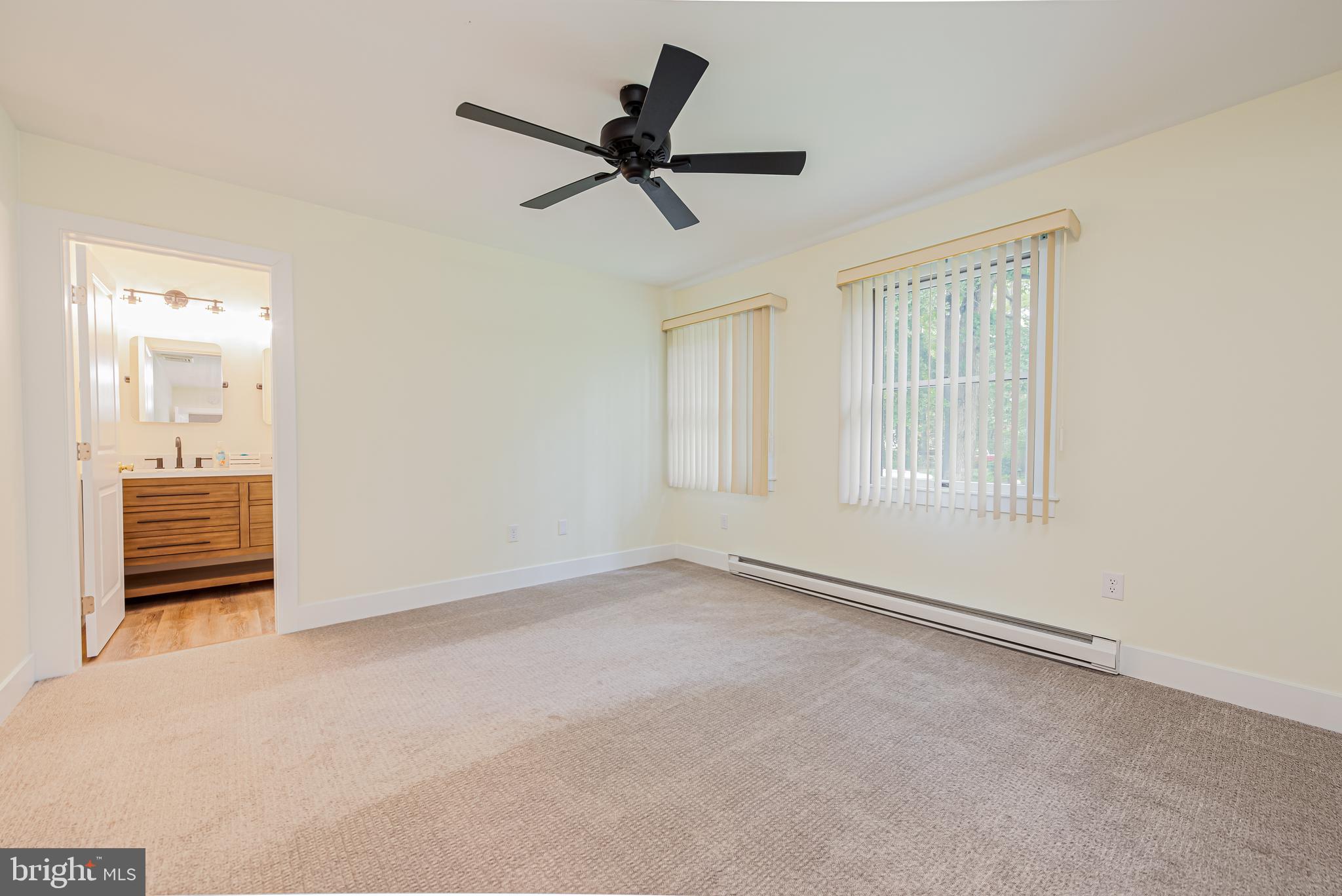 Empty carpeted bedroom with a black ceiling fan, two windows, and an open bathroom doorway.