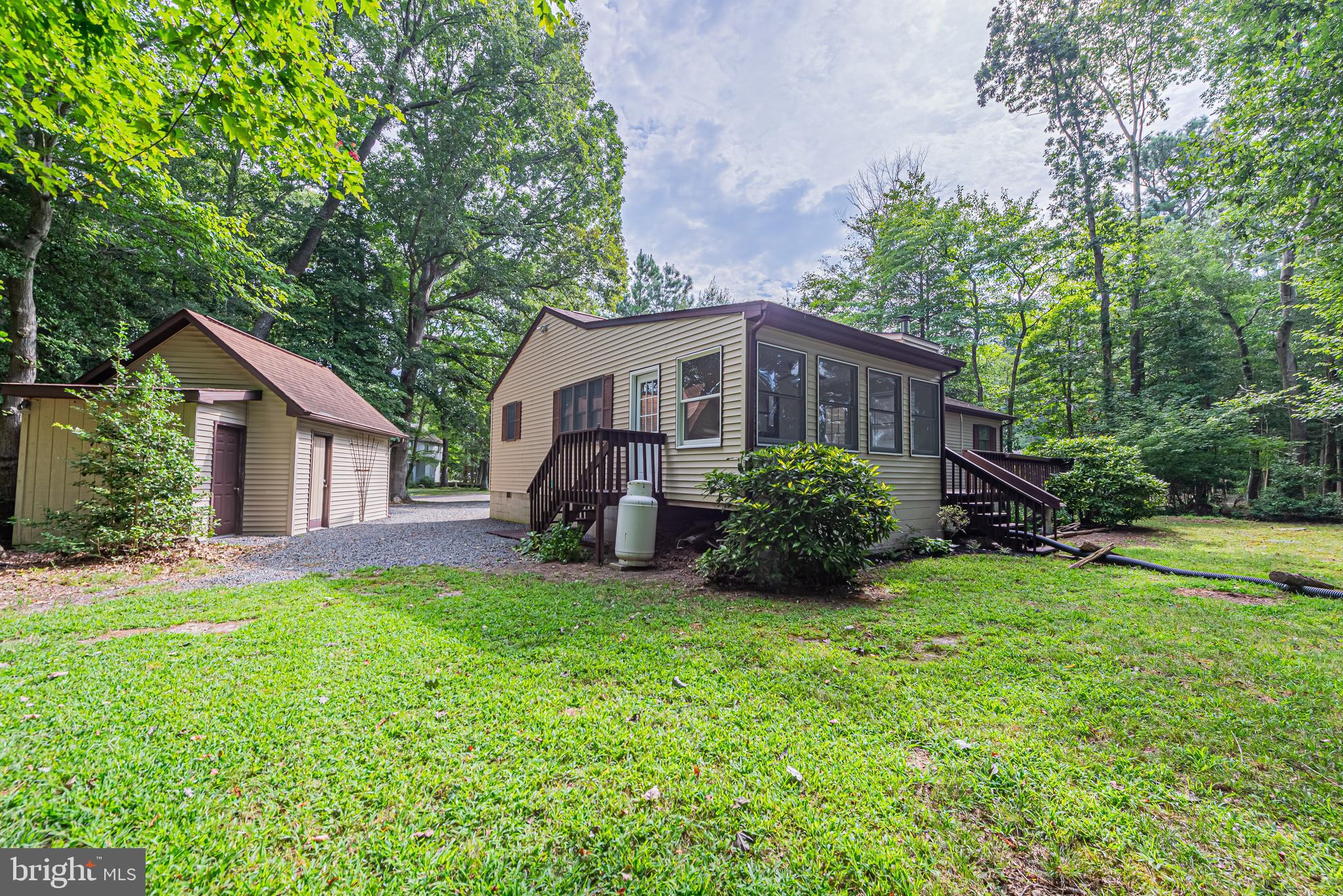 Beige home with a detached outbuilding, gravel driveway, and wooden steps, surrounded by trees.