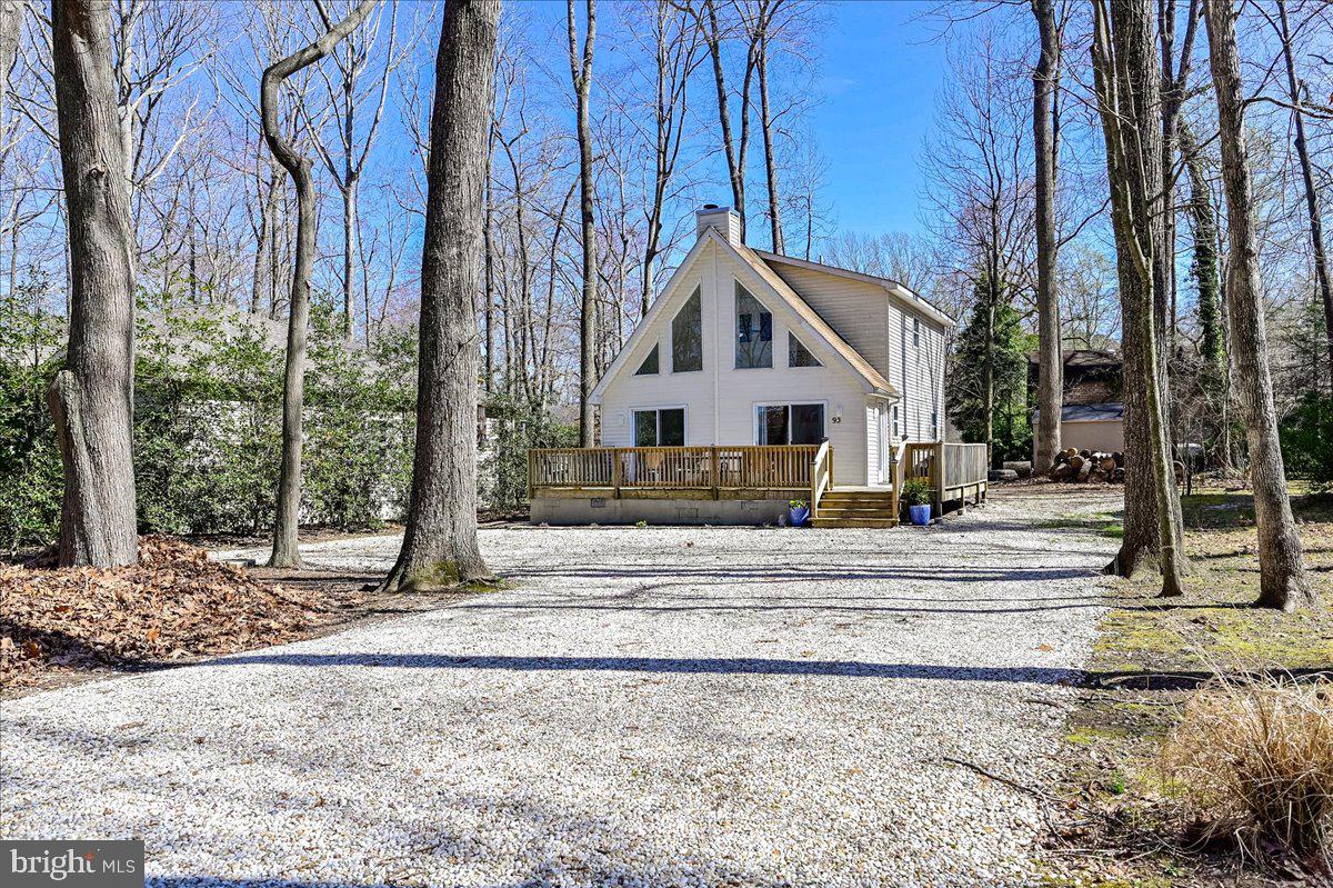 A-frame house with a raised wooden deck among leafless trees beside a gravel driveway.