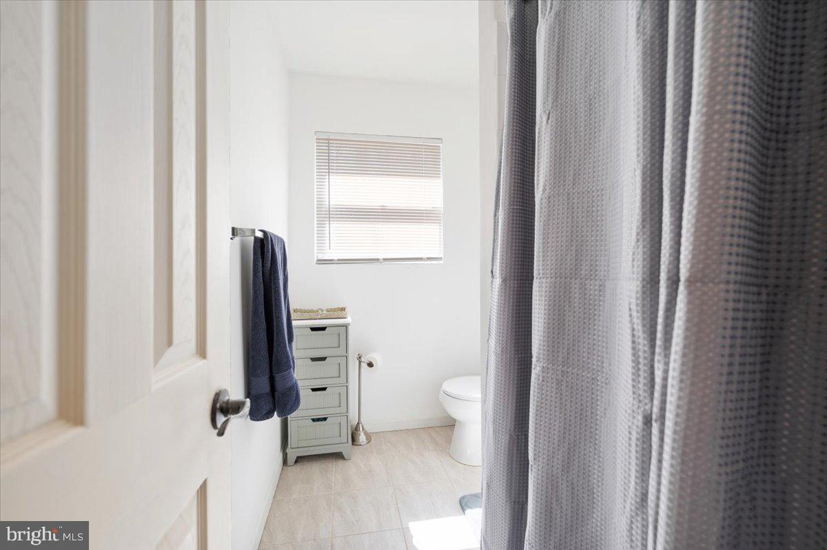 Bathroom with a gray shower curtain, white toilet, and window with blinds.