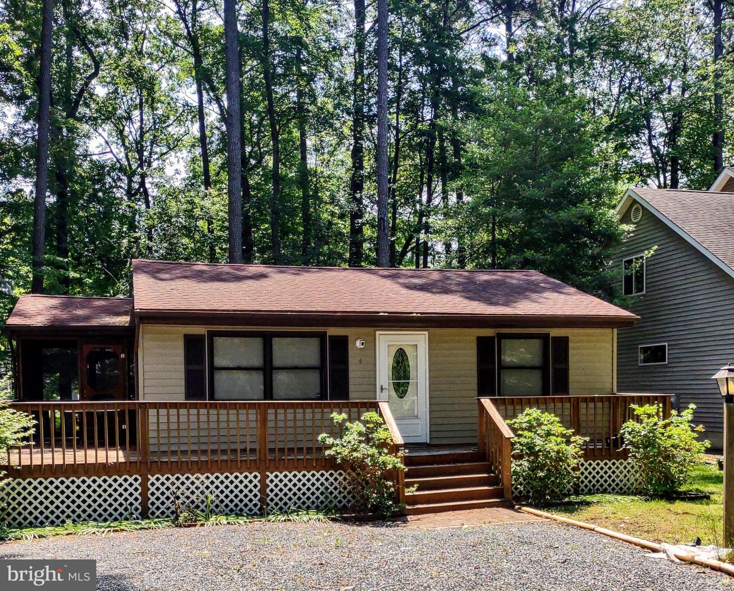 Single-story home with a broad wooden deck and gravel driveway, surrounded by mature trees.