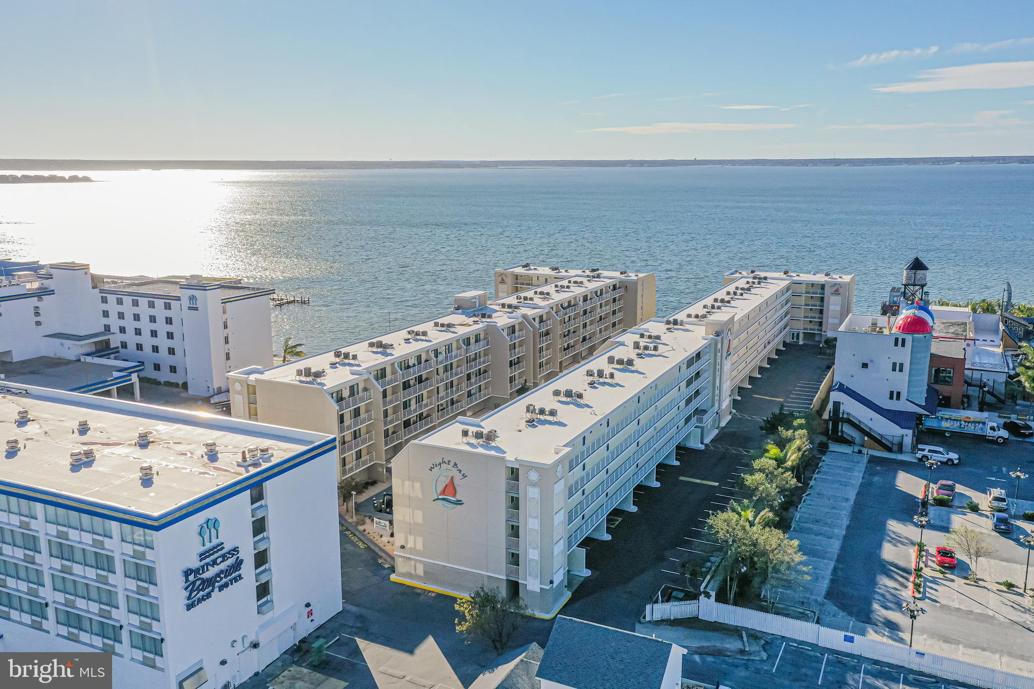Aerial view of waterfront condominium buildings beside the bay, with balconies, parking areas, and neighboring hotels.