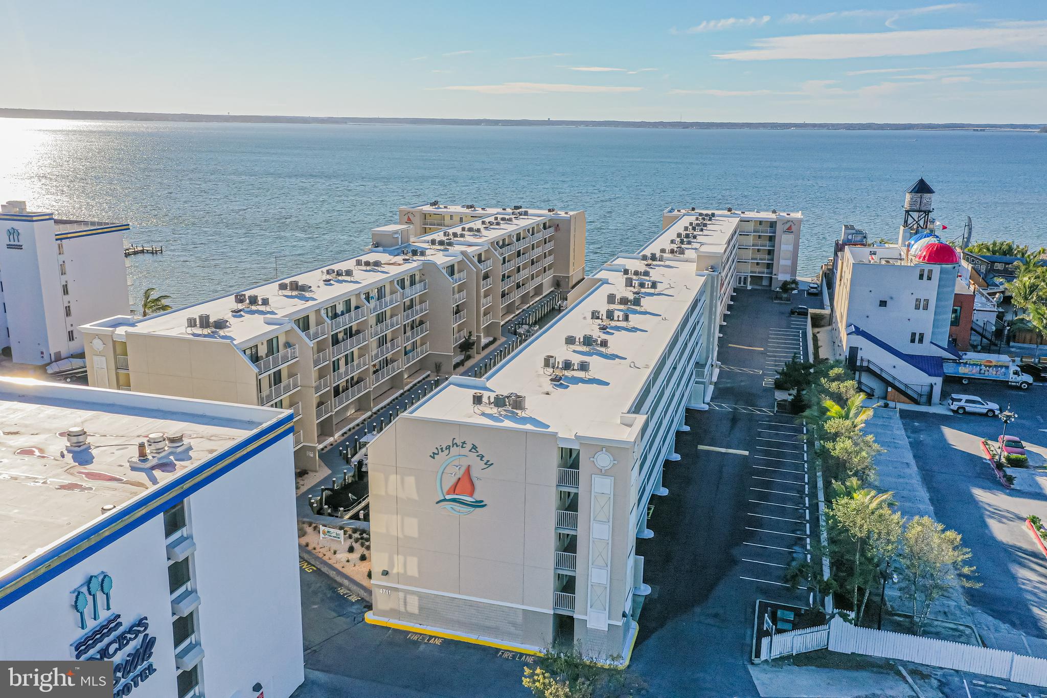 Waterfront condominium buildings with rows of balconies, a parking lot, and open water behind them.