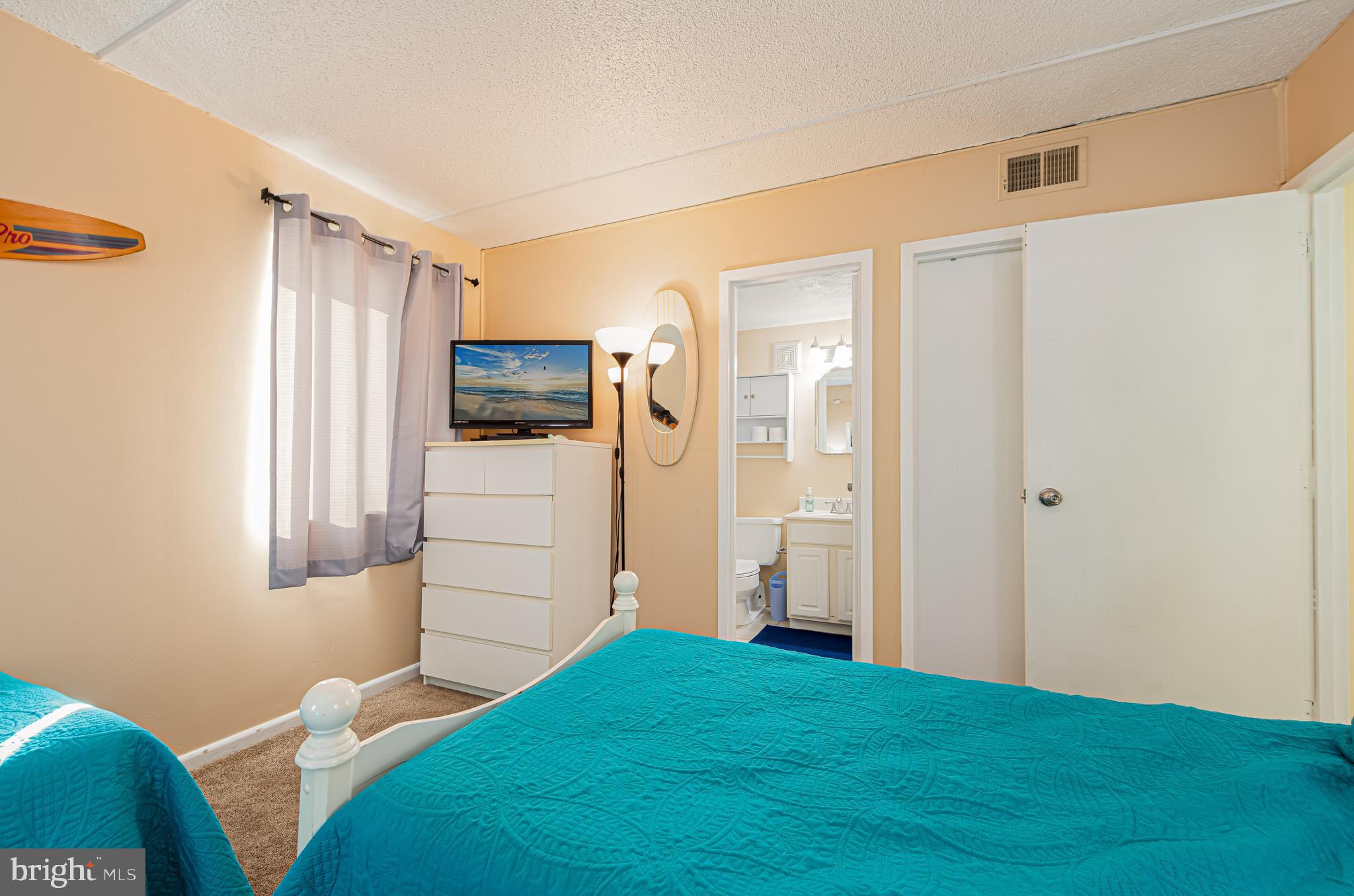 Bedroom with turquoise-covered beds, a white dresser and TV, and an open doorway to the bathroom.