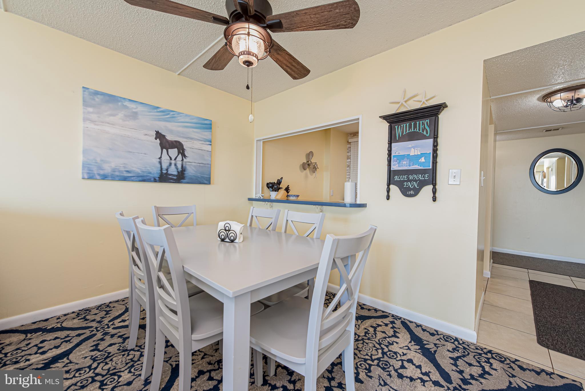 Dining area with a white table and chairs, patterned blue rug, and beach-themed wall decor.