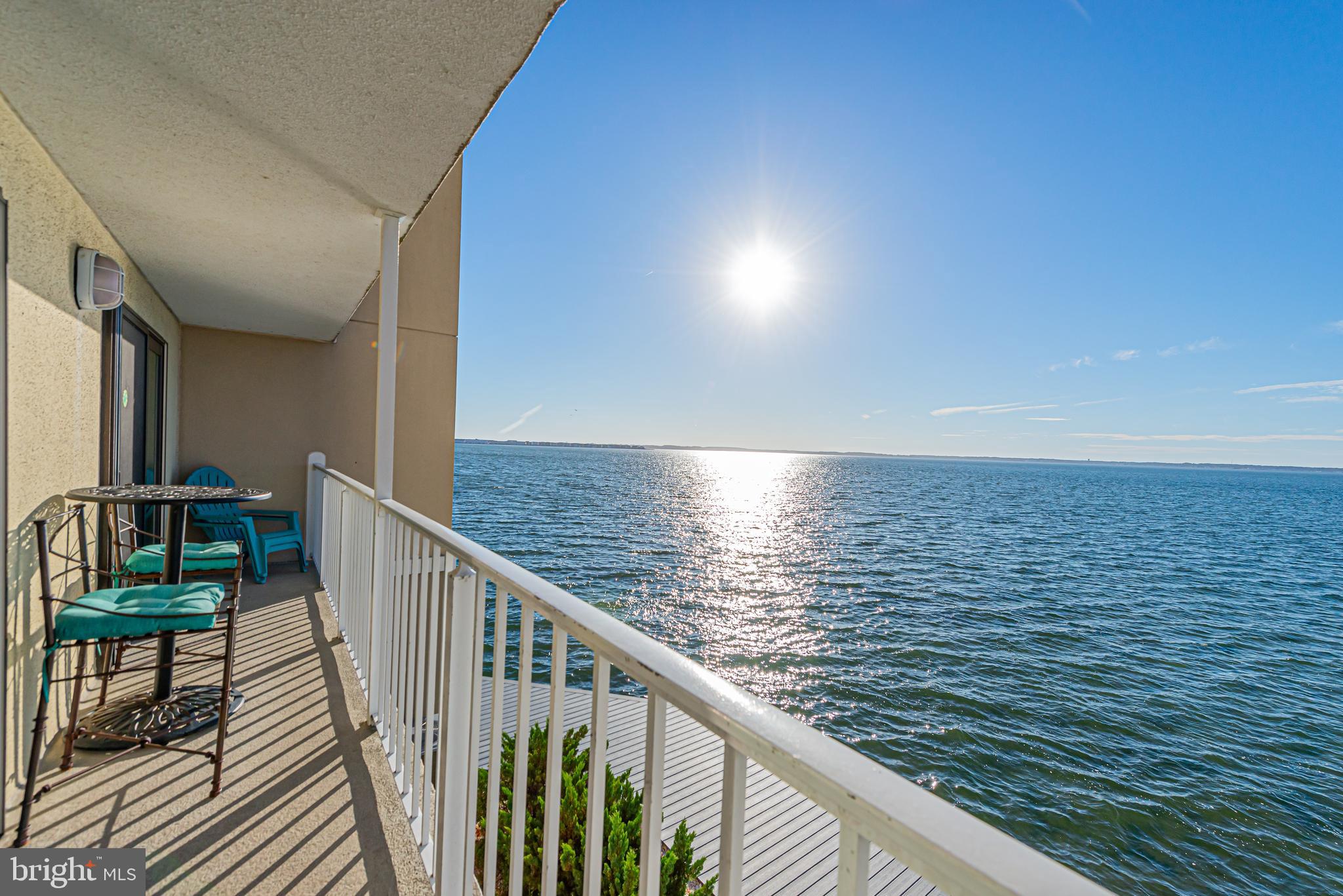 Furnished balcony overlooking sunlit water, with turquoise chairs and a small table.