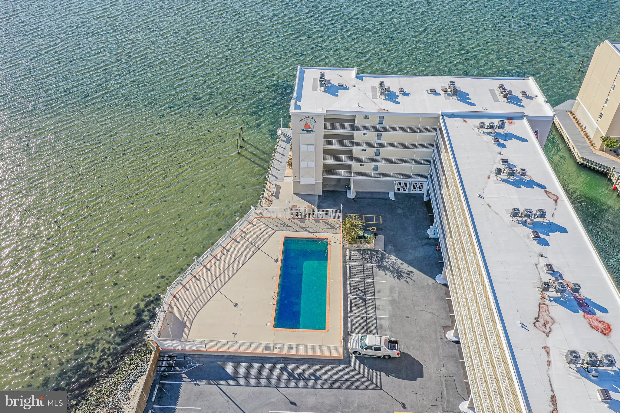 Waterfront condominium building with an outdoor pool beside green-blue water, viewed from above.