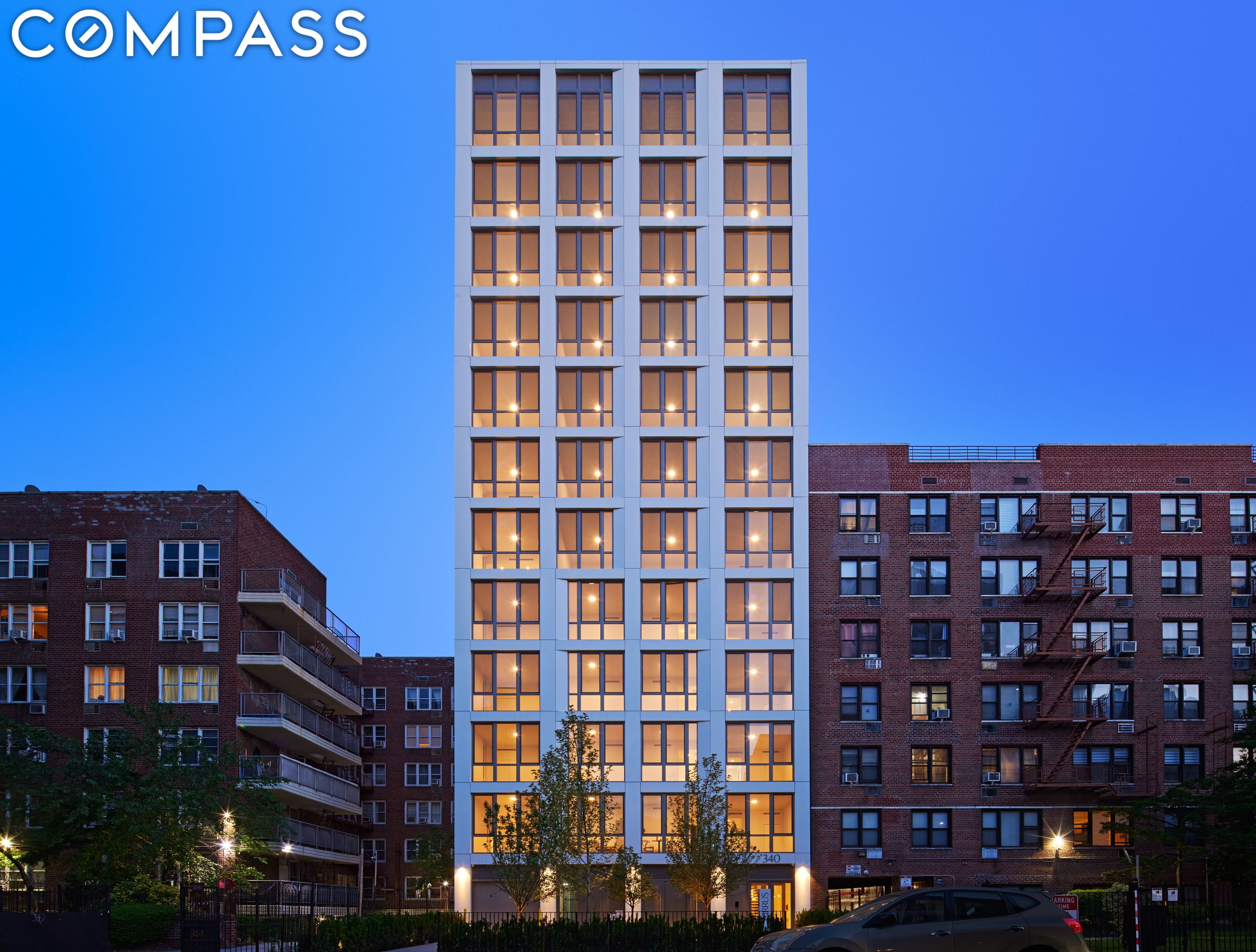 Modern white apartment building with illuminated windows between brick buildings at dusk.