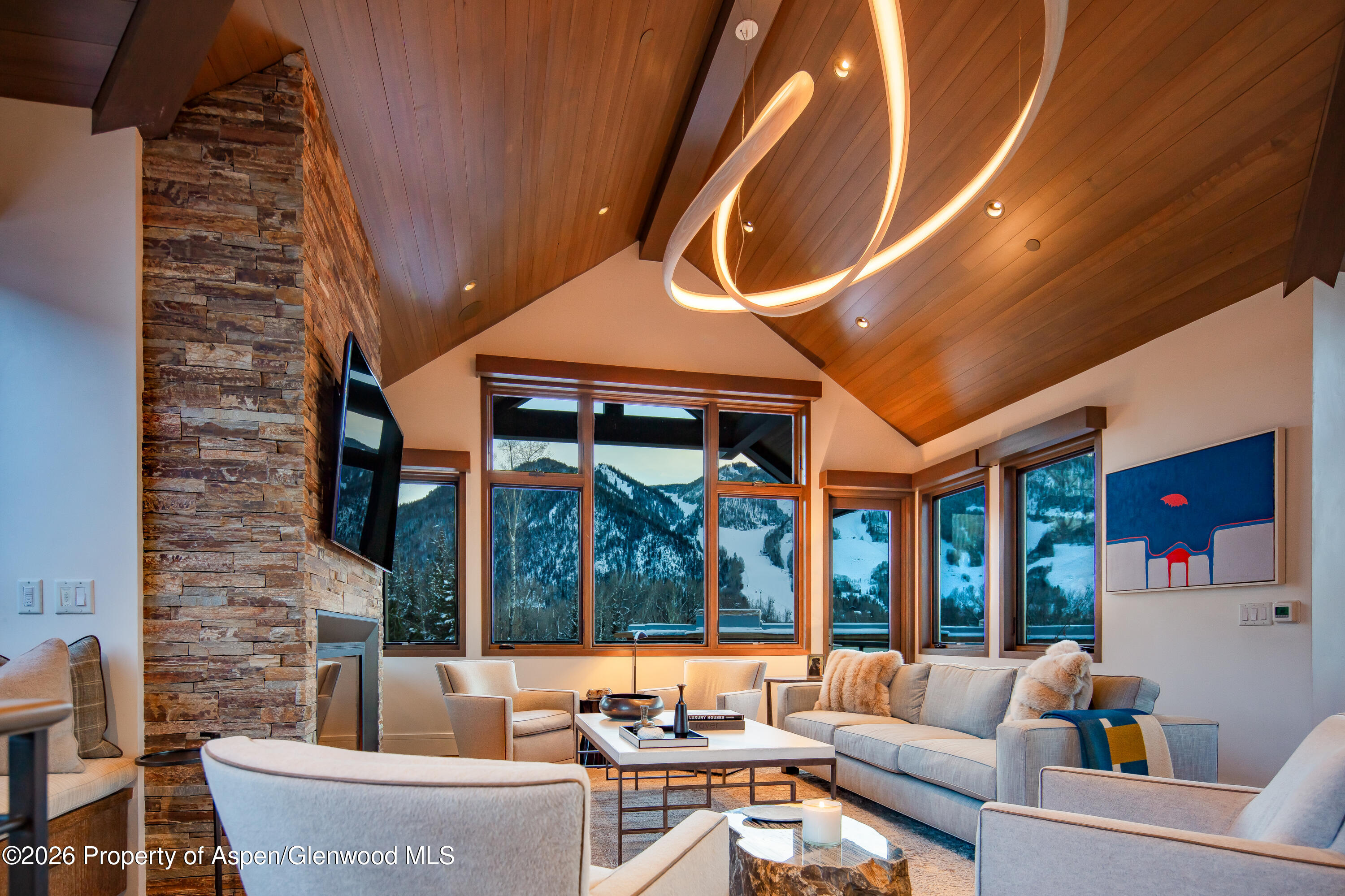 Vaulted living room with a stone fireplace, sculptural chandelier, and snowy mountain views through large windows.