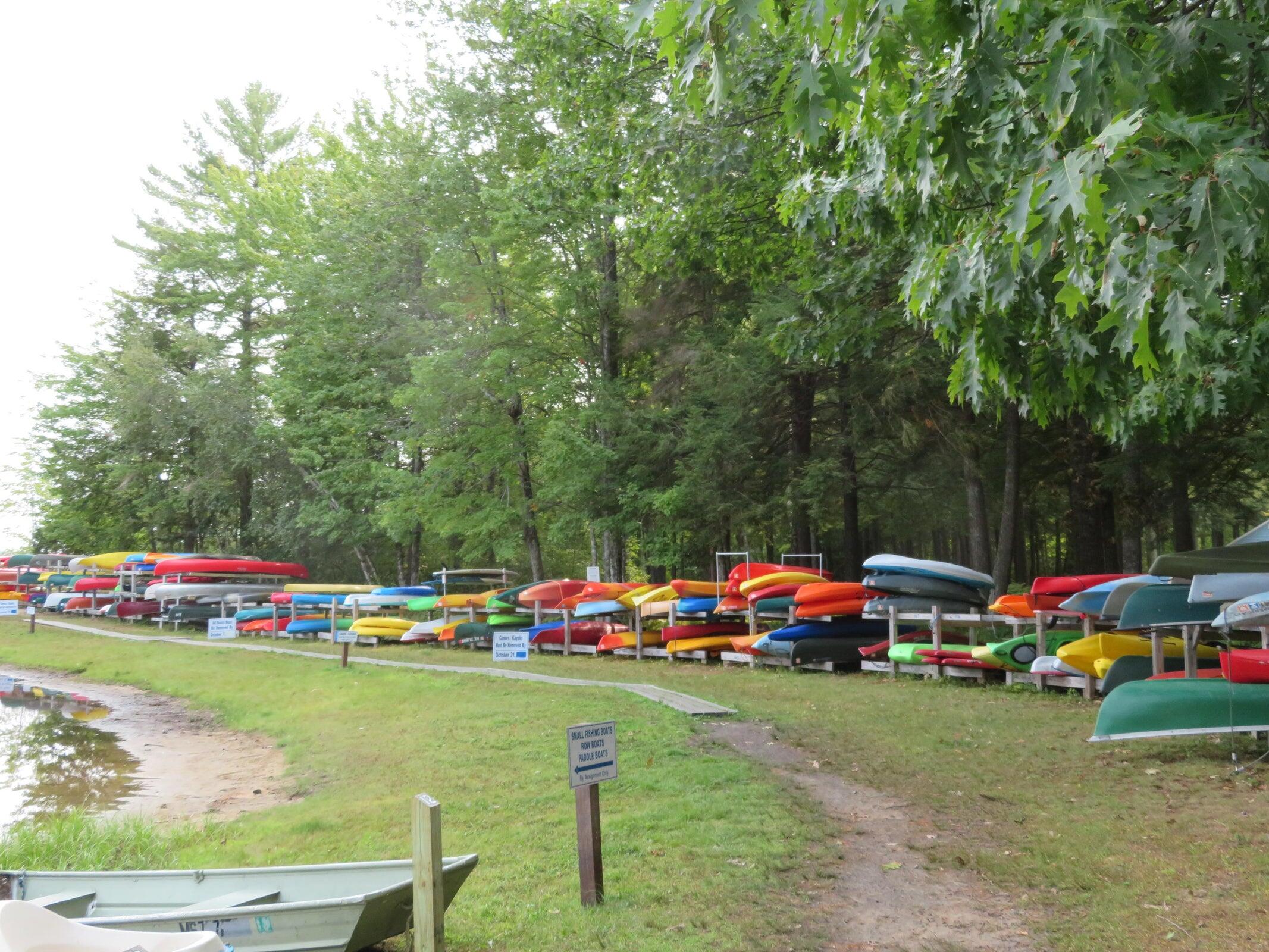 Colorful kayaks and small boats line racks beside a wooded shoreline and grassy path.