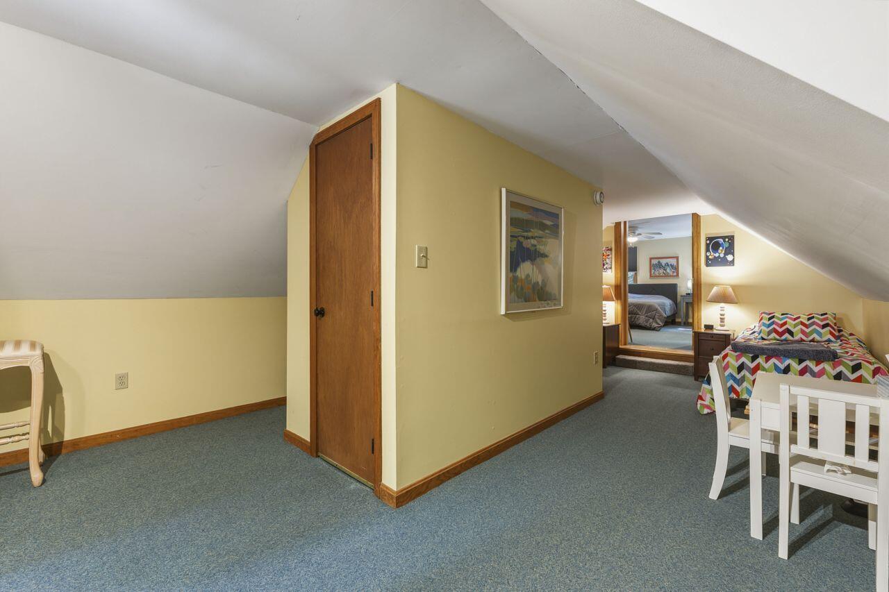 Attic bedroom with sloped ceilings, blue carpet, and a colorful bed beside white furniture.