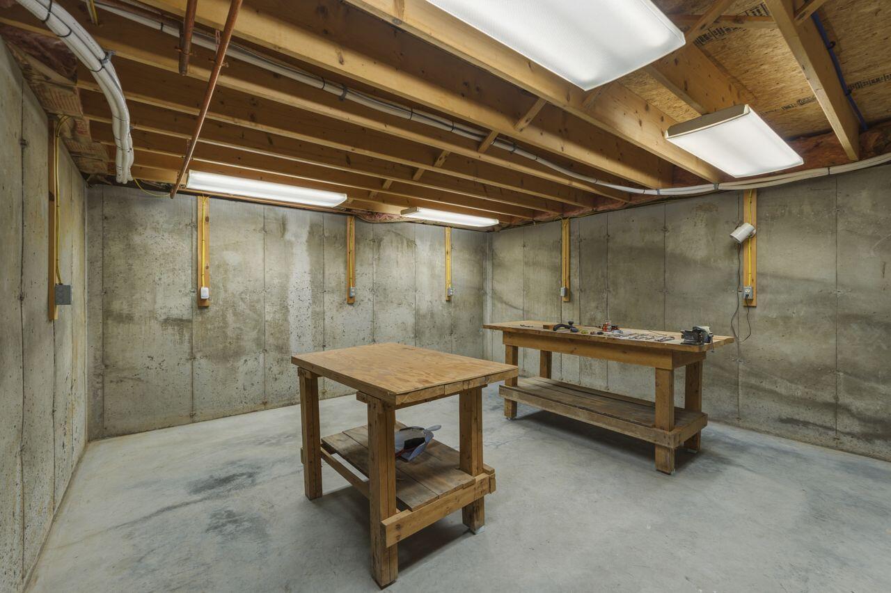 Basement workshop with two wooden workbenches, exposed ceiling joists, and concrete walls.