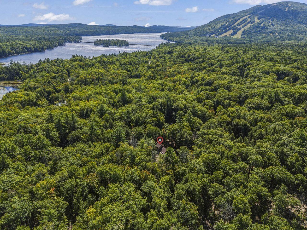 Aerial view of a forest-surrounded home with a lake and wooded hills in the distance.