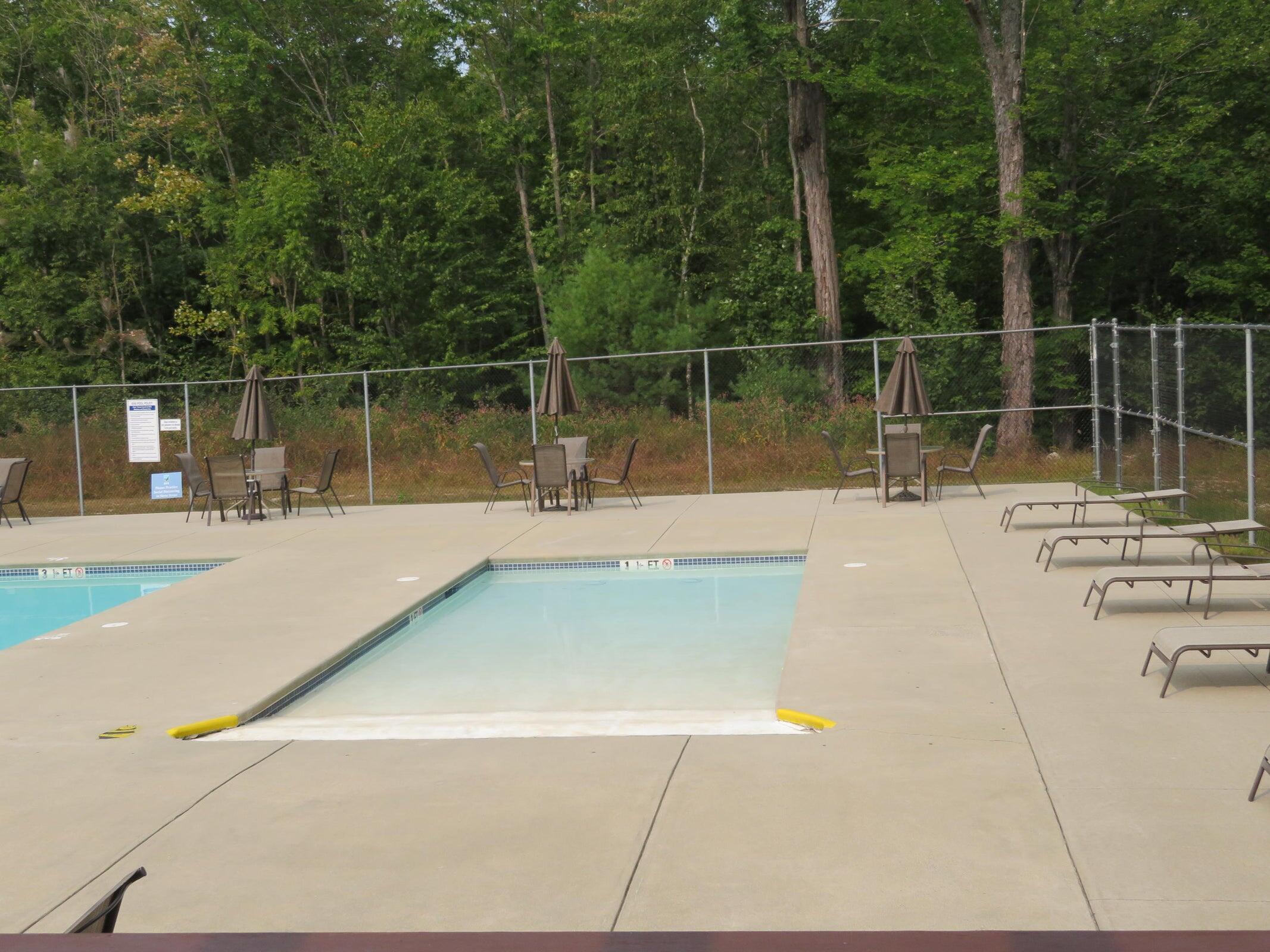 Fenced outdoor pool with a shallow-entry section, patio seating, and lounge chairs beside woods.