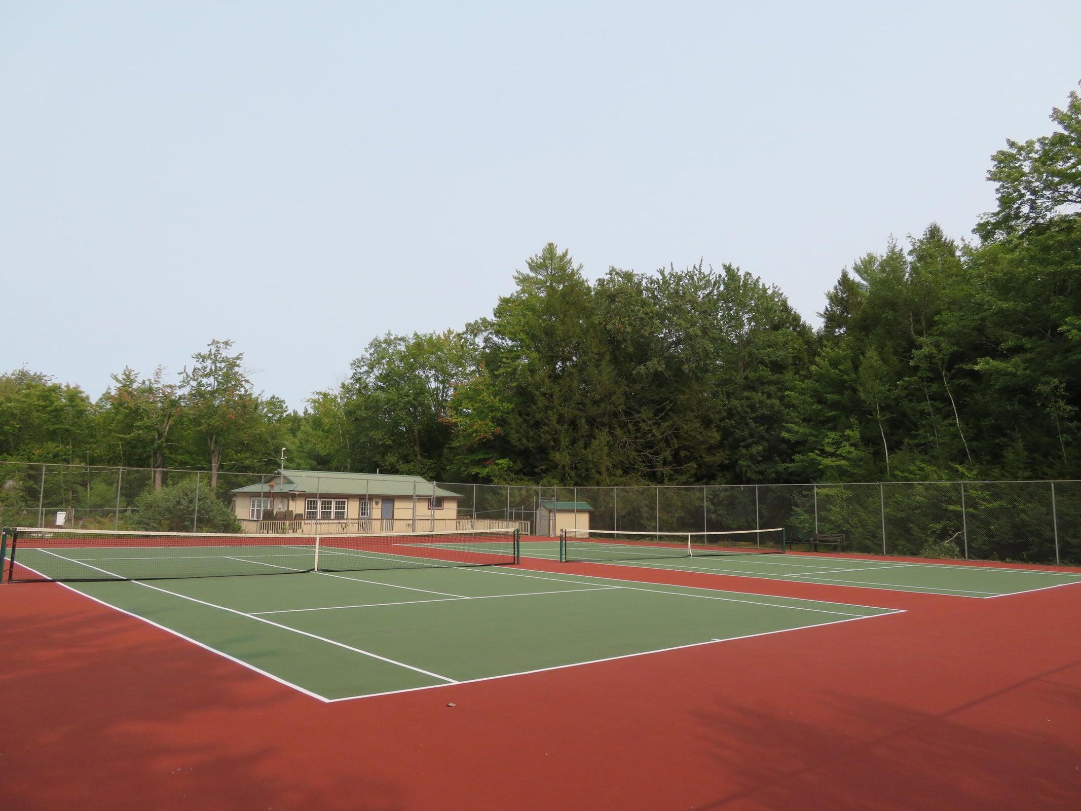 Fenced tennis courts with green playing surfaces and red surrounds, backed by a small clubhouse and woods.