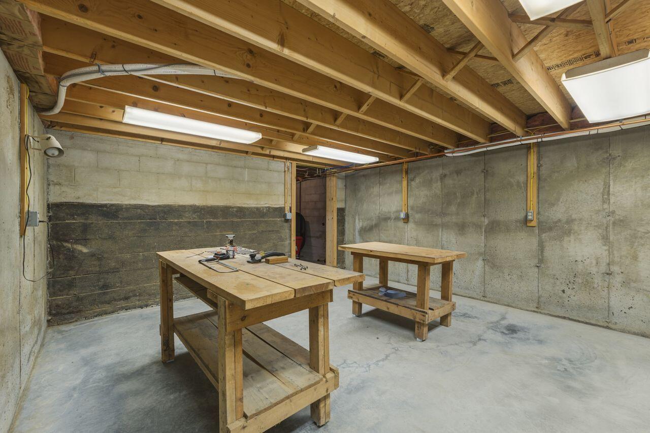 Basement workshop with wooden workbenches, exposed joists, and concrete walls.