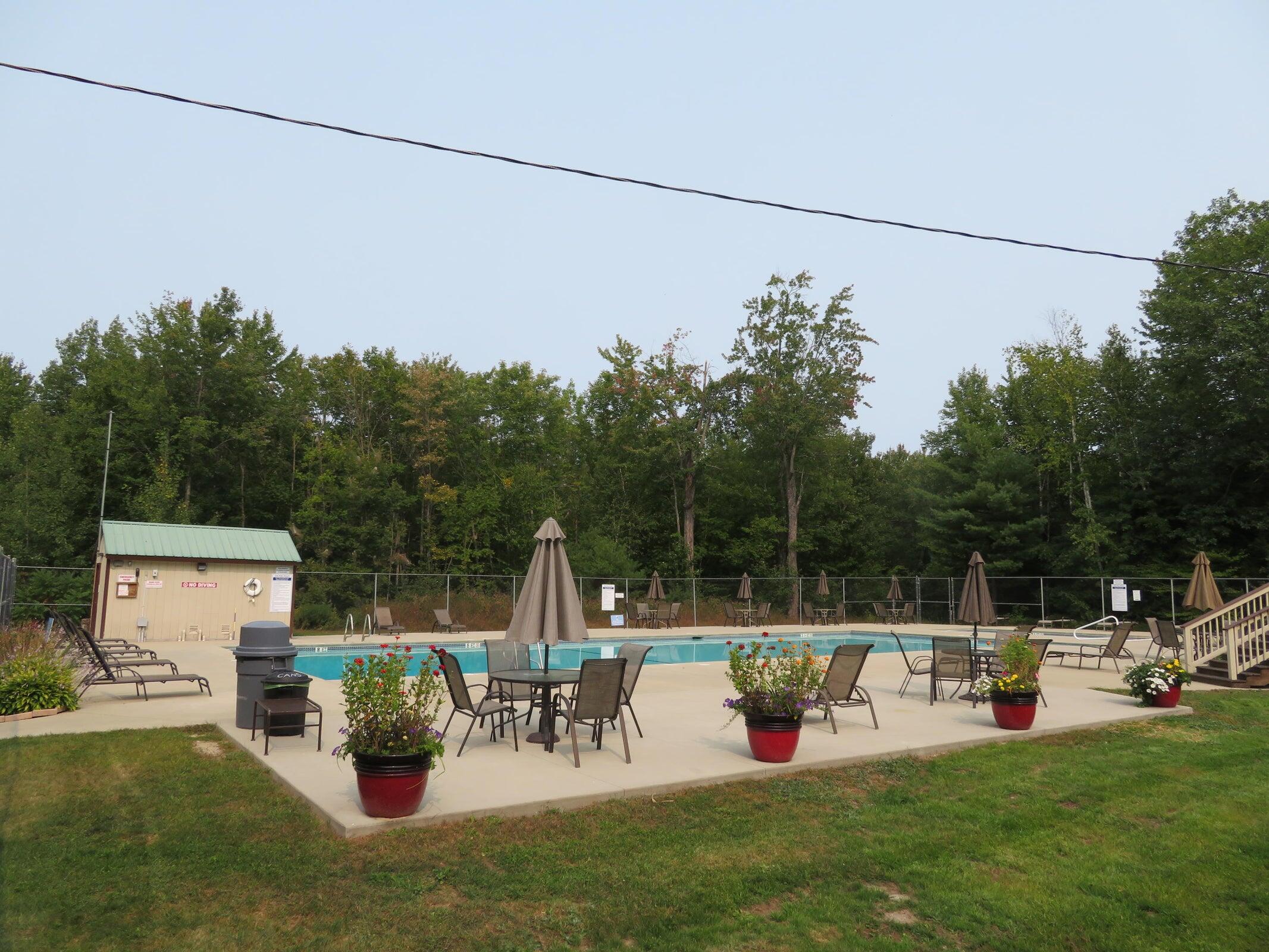 Outdoor pool surrounded by patio seating, closed umbrellas, potted flowers, and trees.