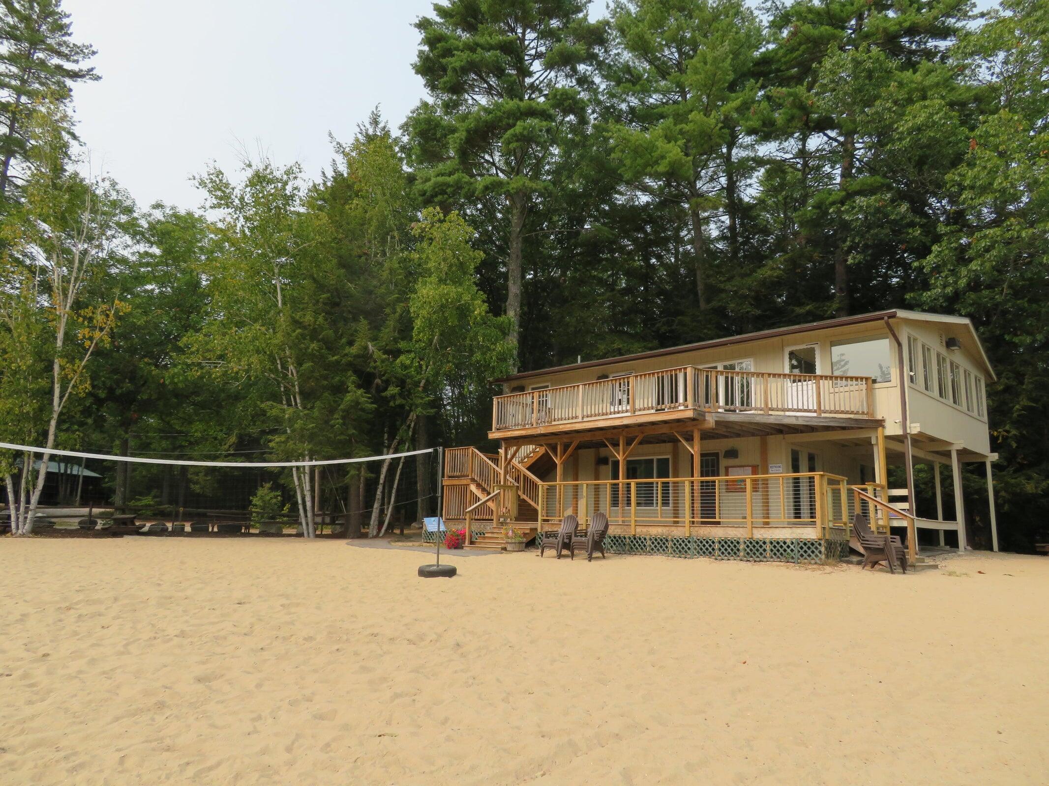 Raised tan house with broad decks behind a sandy beach and volleyball net, surrounded by mature trees.