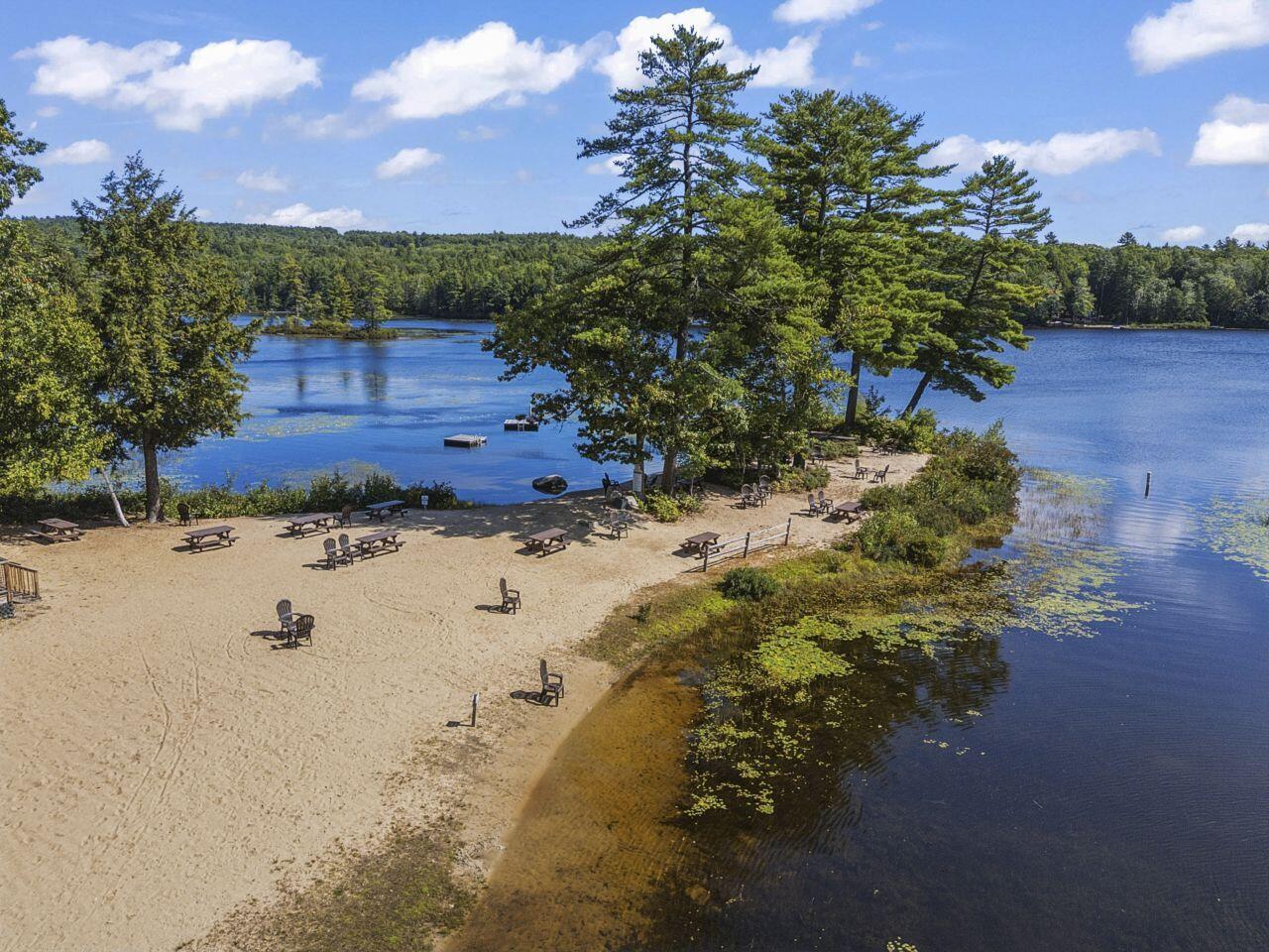 Aerial view of a sandy lakeside beach with picnic tables, chairs, and wooded shoreline.