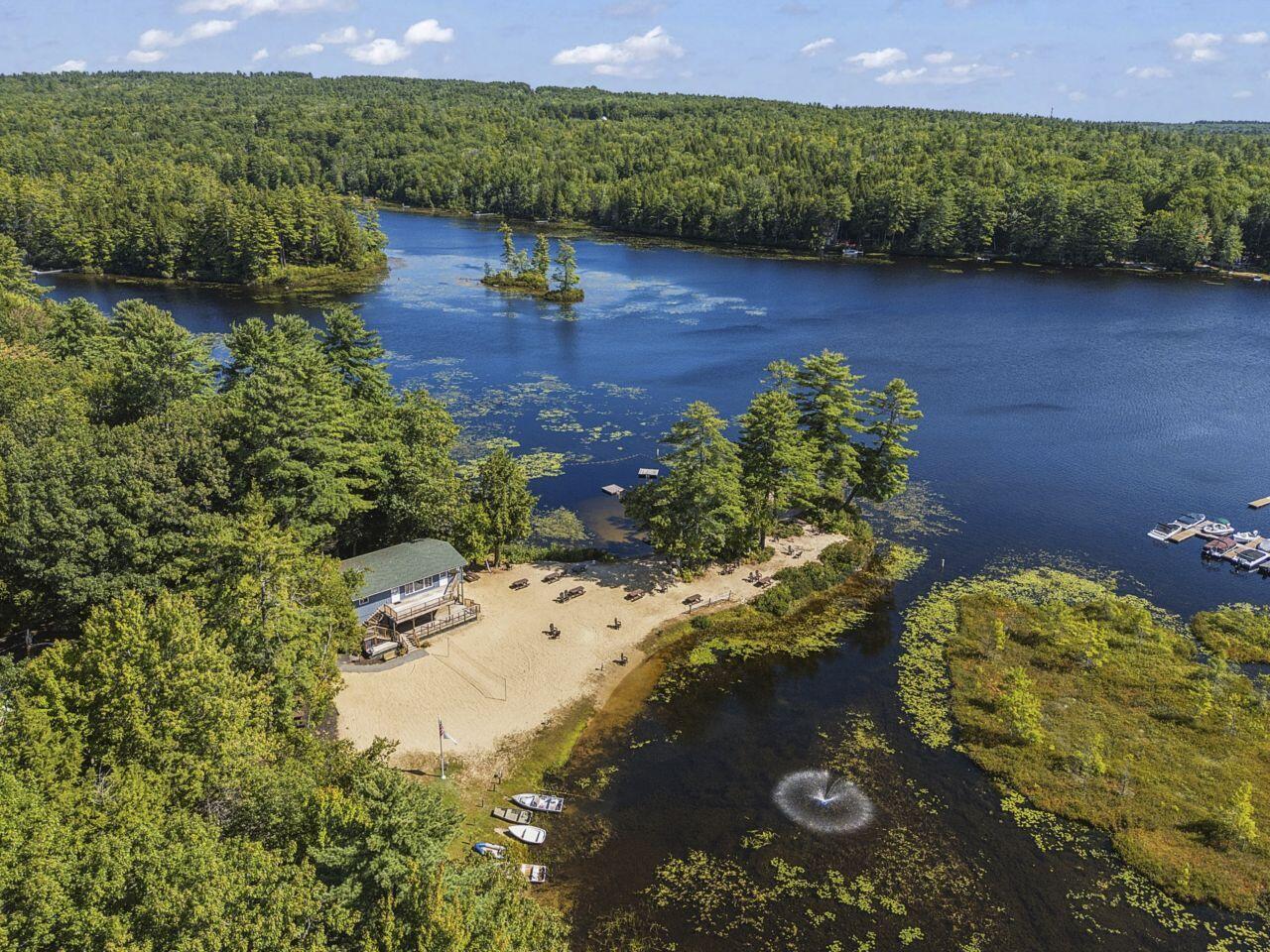 Aerial view of a wooded lakefront property with a sandy beach and fountain in the water.