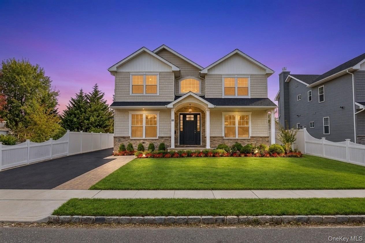 Two-story gray home with glowing windows, landscaped lawn, white fencing, and paved driveway at dusk.