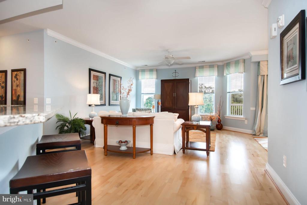 Living room with a white sofa, round wooden table, hardwood floors, and tall windows overlooking greenery.