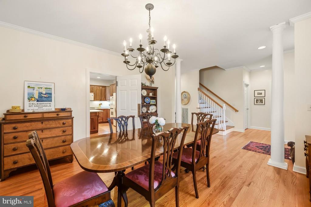 Dining room with a long wooden table, ornate chandelier, hardwood floors, and an open view to the kitchen and staircase.