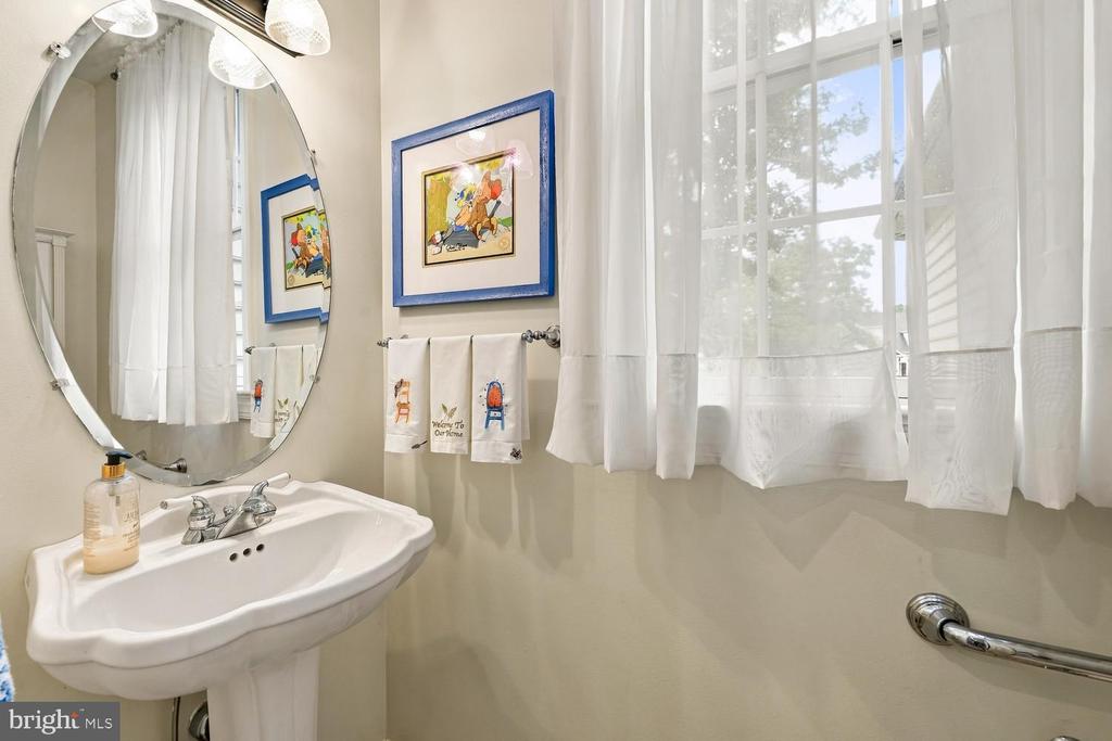 Bathroom with a pedestal sink, large oval mirror, sheer-curtained window, and illustrated towels.