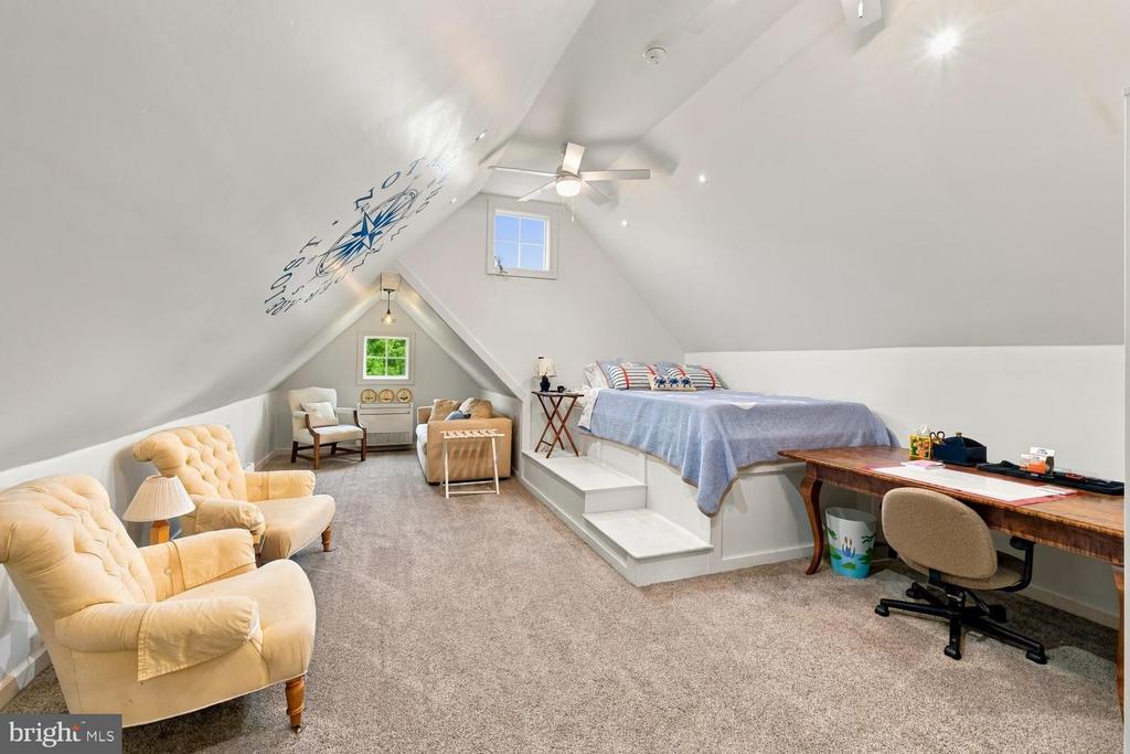 Furnished attic loft bedroom with a raised bed platform, desk, and seating area beneath a vaulted ceiling.