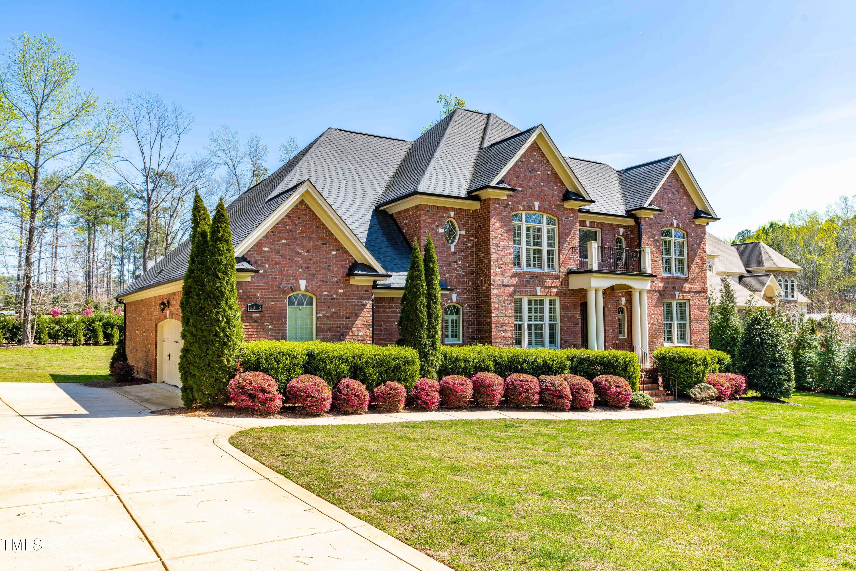 Two-story red-brick home with a balcony, pink flowering shrubs, and a curved driveway.