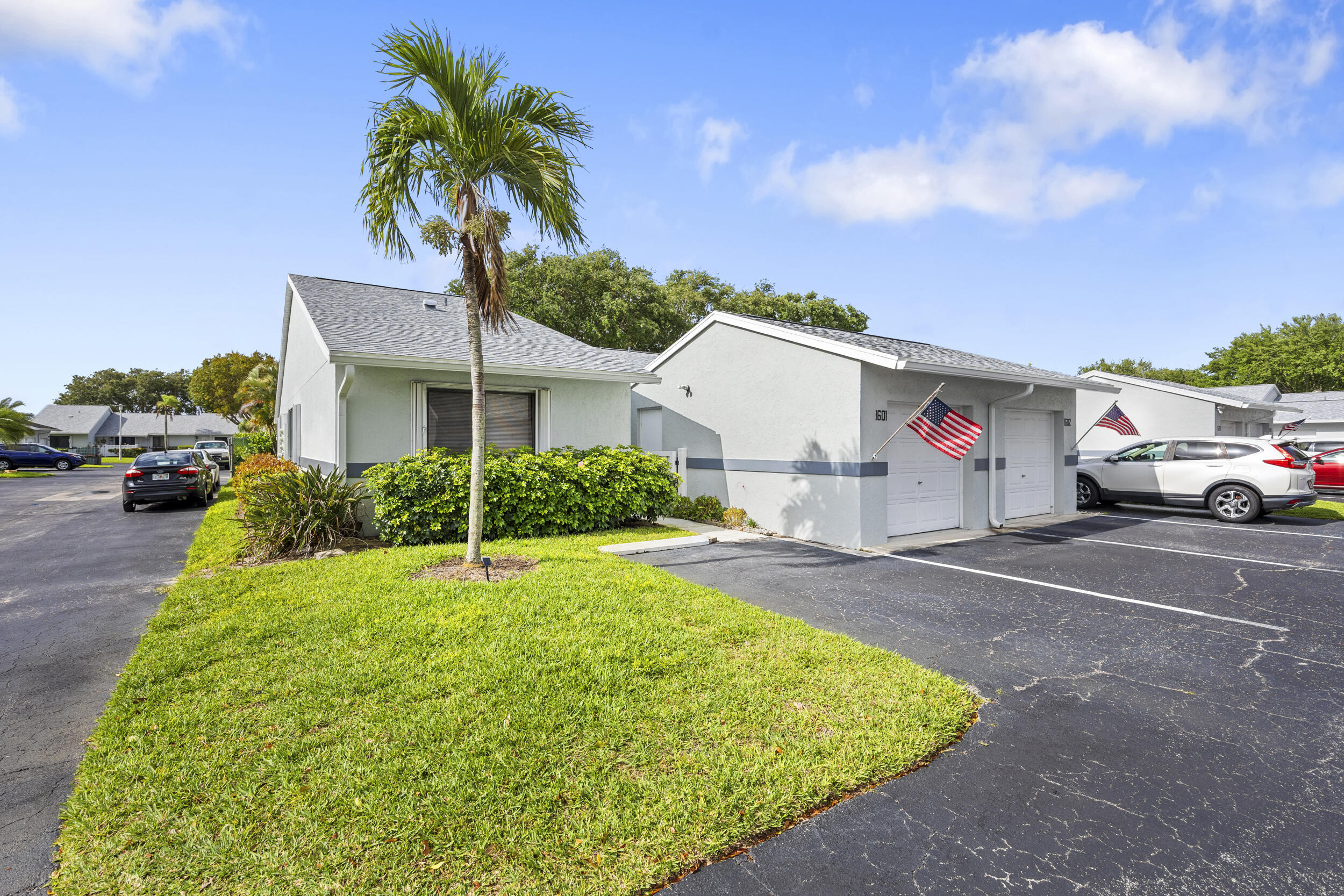 Single-story white condominium with a palm tree, landscaped lawn, parking, and American flags by the entrances.