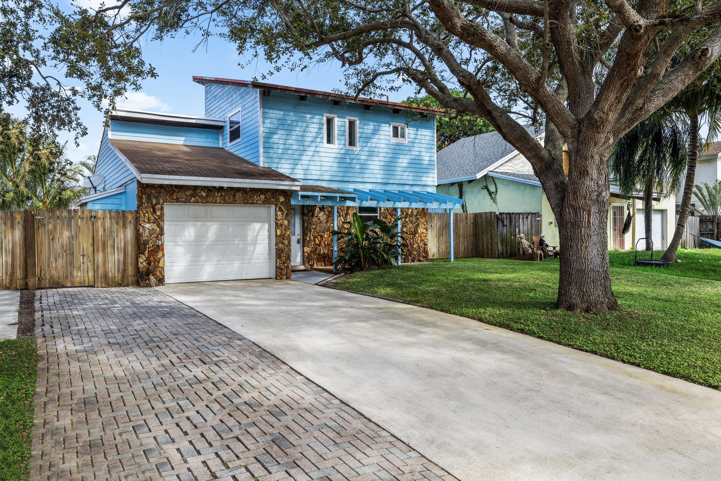 Blue two-story home with a stone facade, paved driveway, and large oak tree in the front yard.