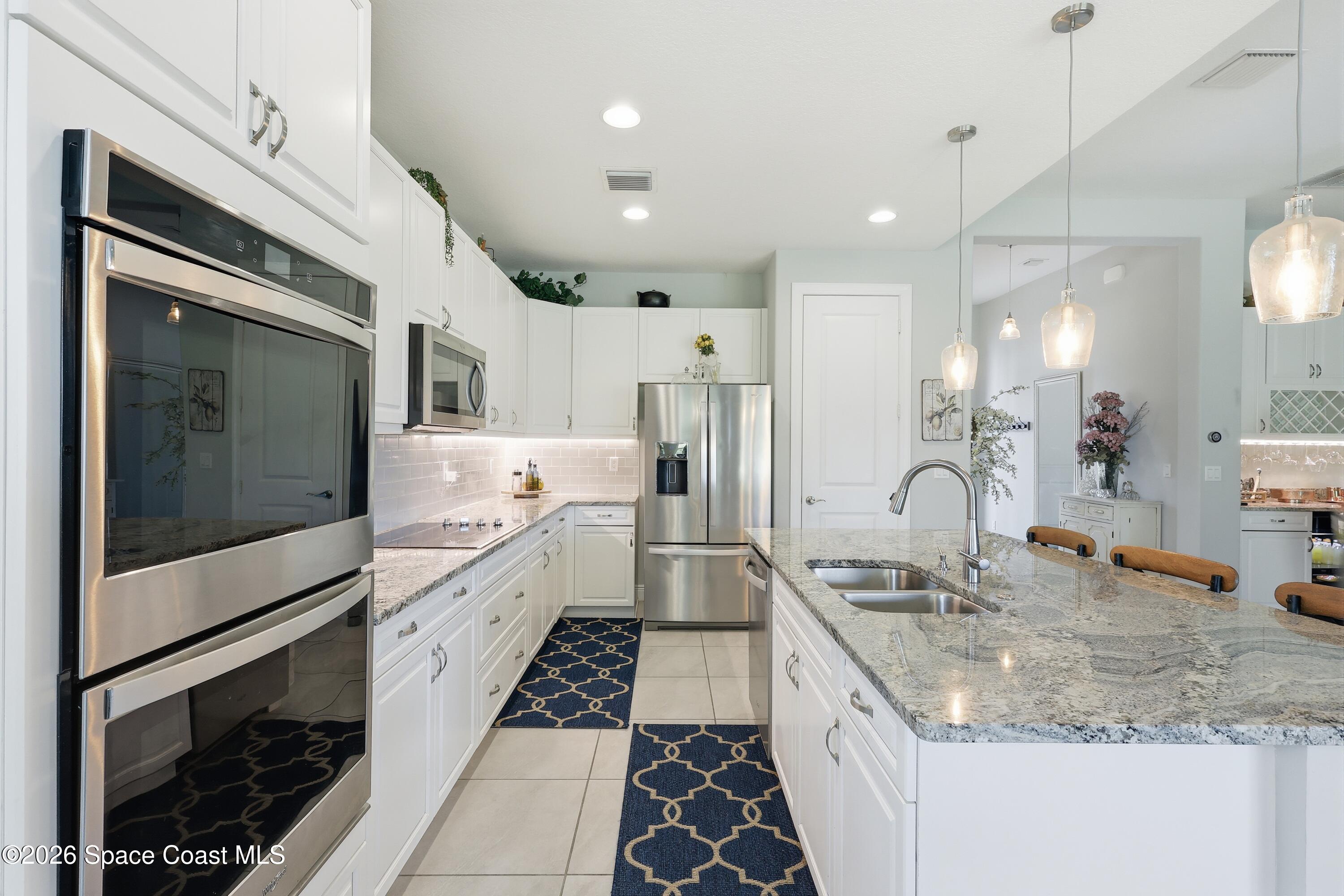 Bright white kitchen with stainless steel appliances, granite island, double sink, and navy patterned runners.
