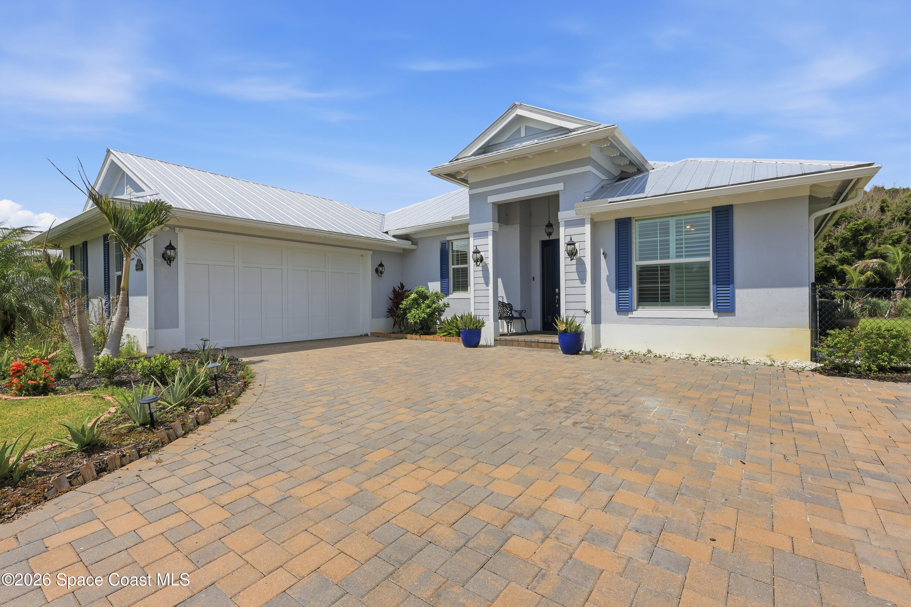 House front with blue shutters, white metal roof, tropical landscaping, and a wide paver driveway.