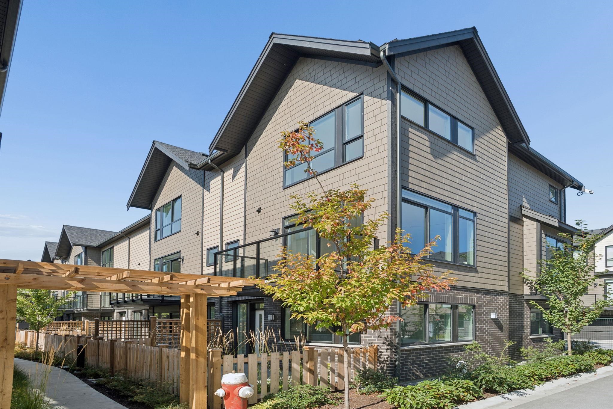 Modern townhouses with gray siding, fenced patios, a wooden pergola, and landscaped trees.