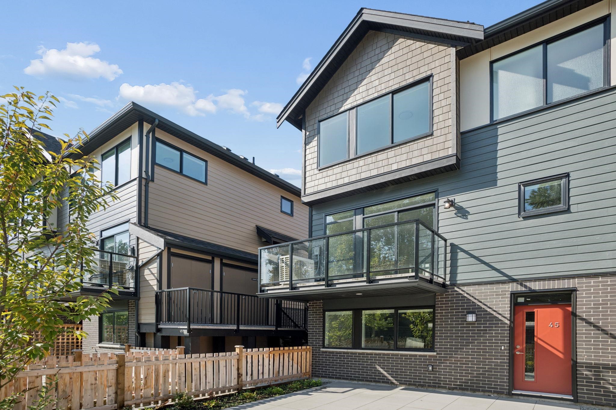 Modern attached townhouses with glass balconies, fenced patios, and a red entrance door.