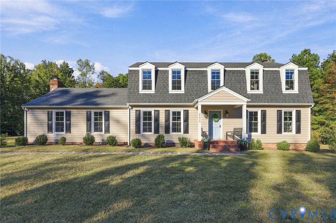 Beige house with five roof dormers, black shutters, and a covered entrance facing a broad lawn.