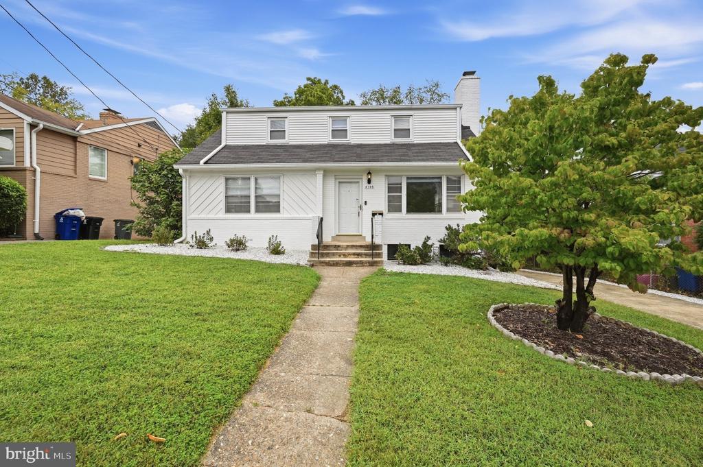 White two-story house with a front walkway, broad lawn, and leafy tree.