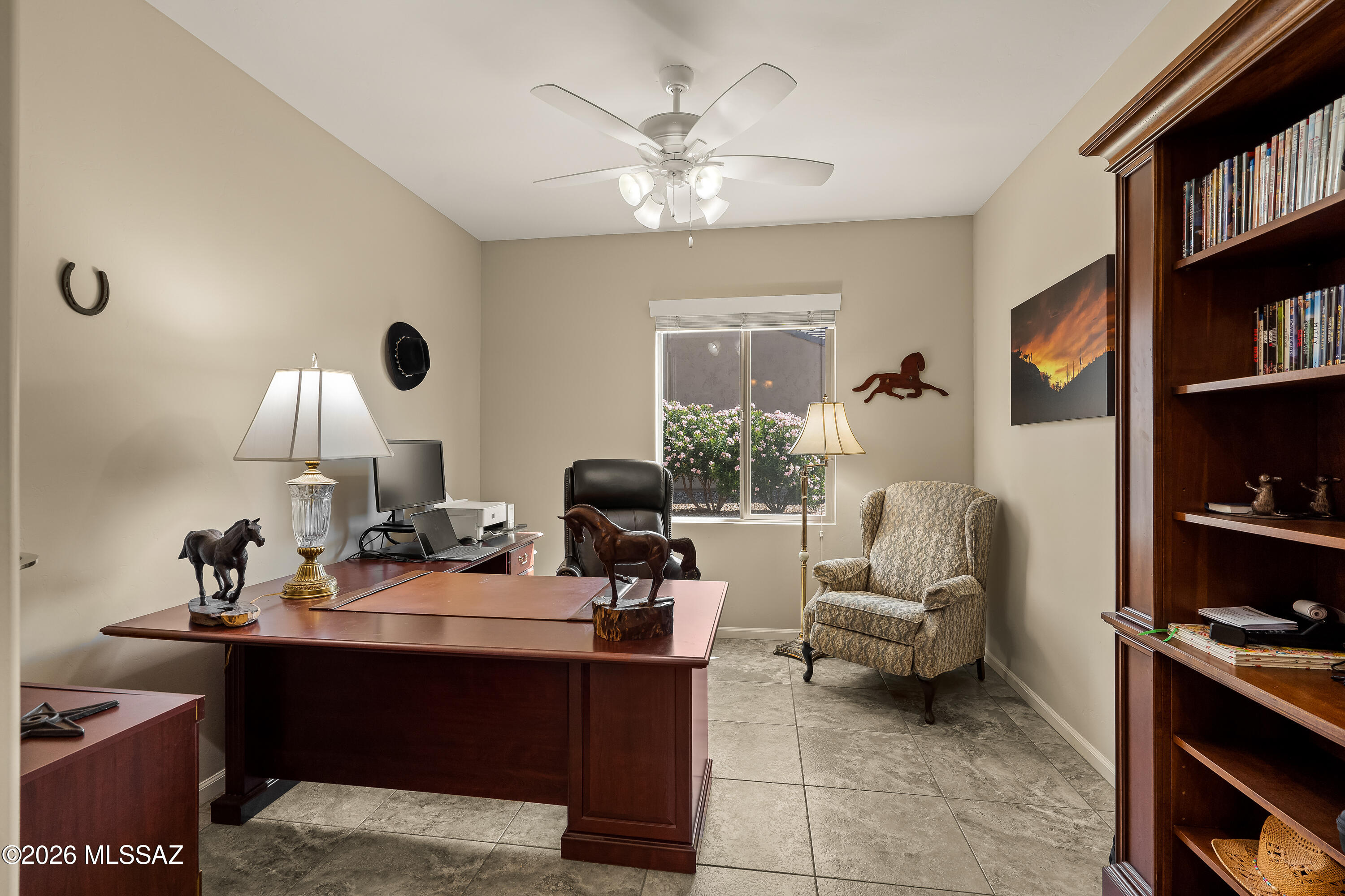 Furnished home office with a large wood desk, built-in bookcase, and window overlooking flowering shrubs.