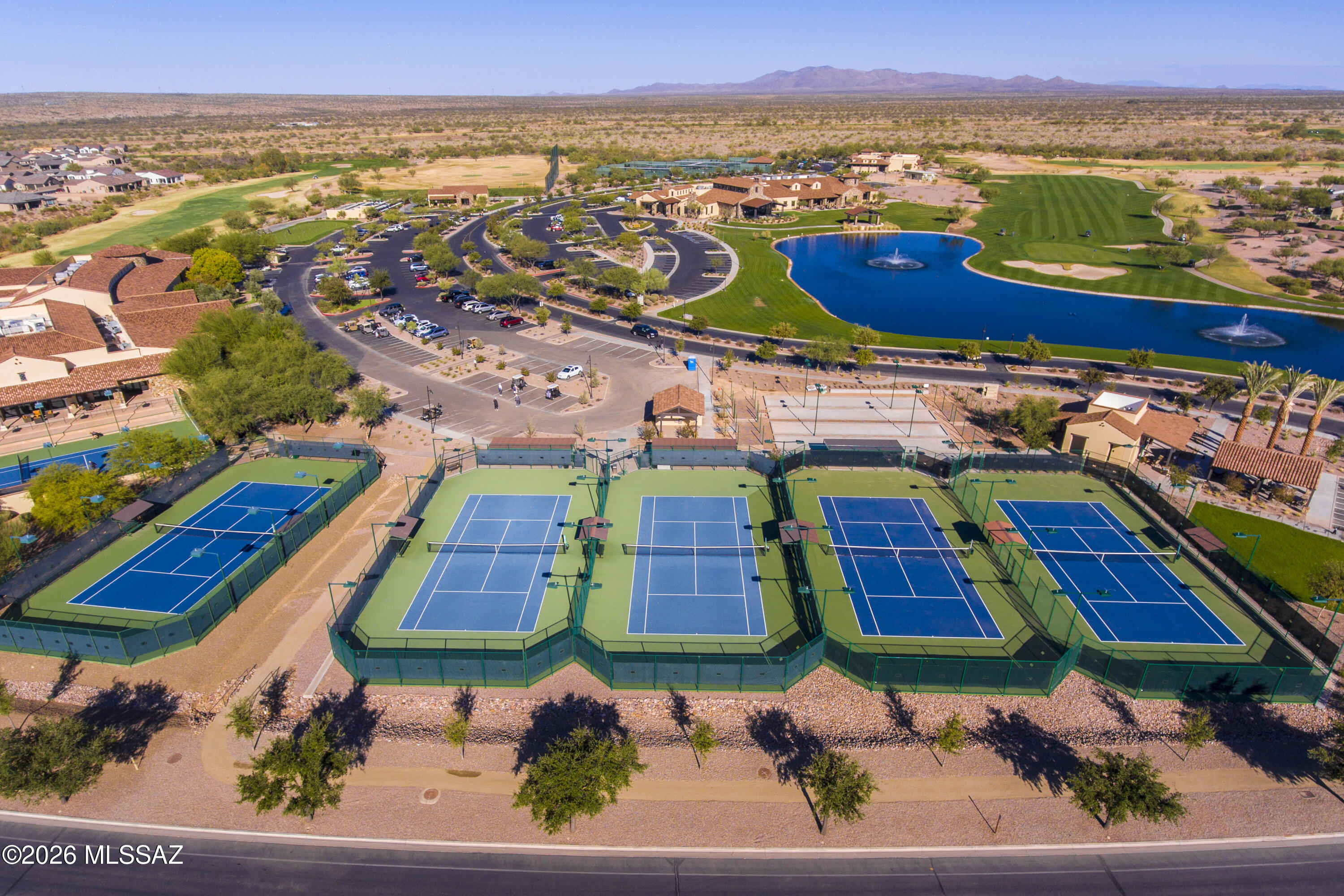 Aerial view of blue courts beside a golf course, fountain lake, and clubhouse in the desert.