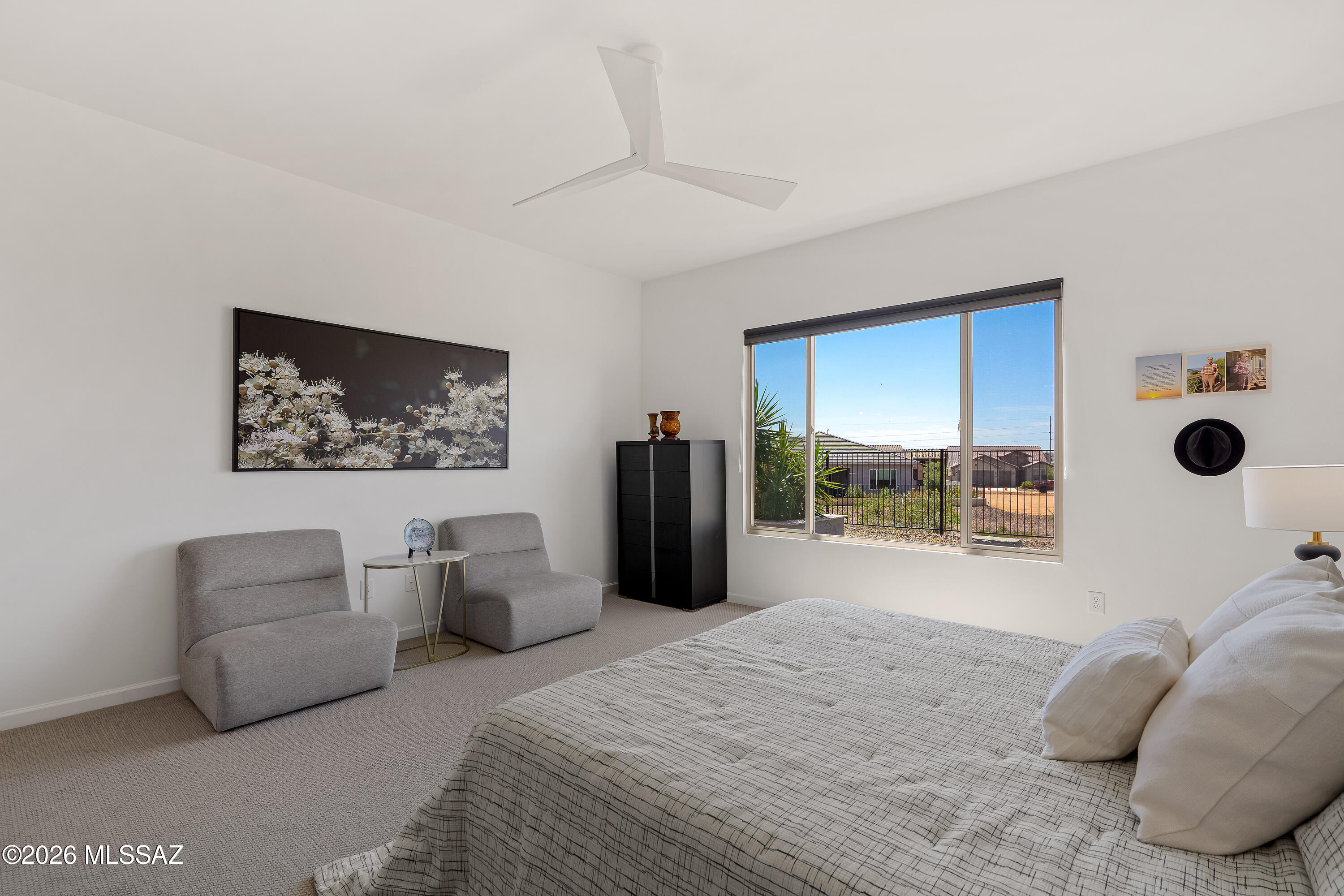 Furnished bedroom with a large bed, gray lounge chairs, and a wide window overlooking neighboring homes.