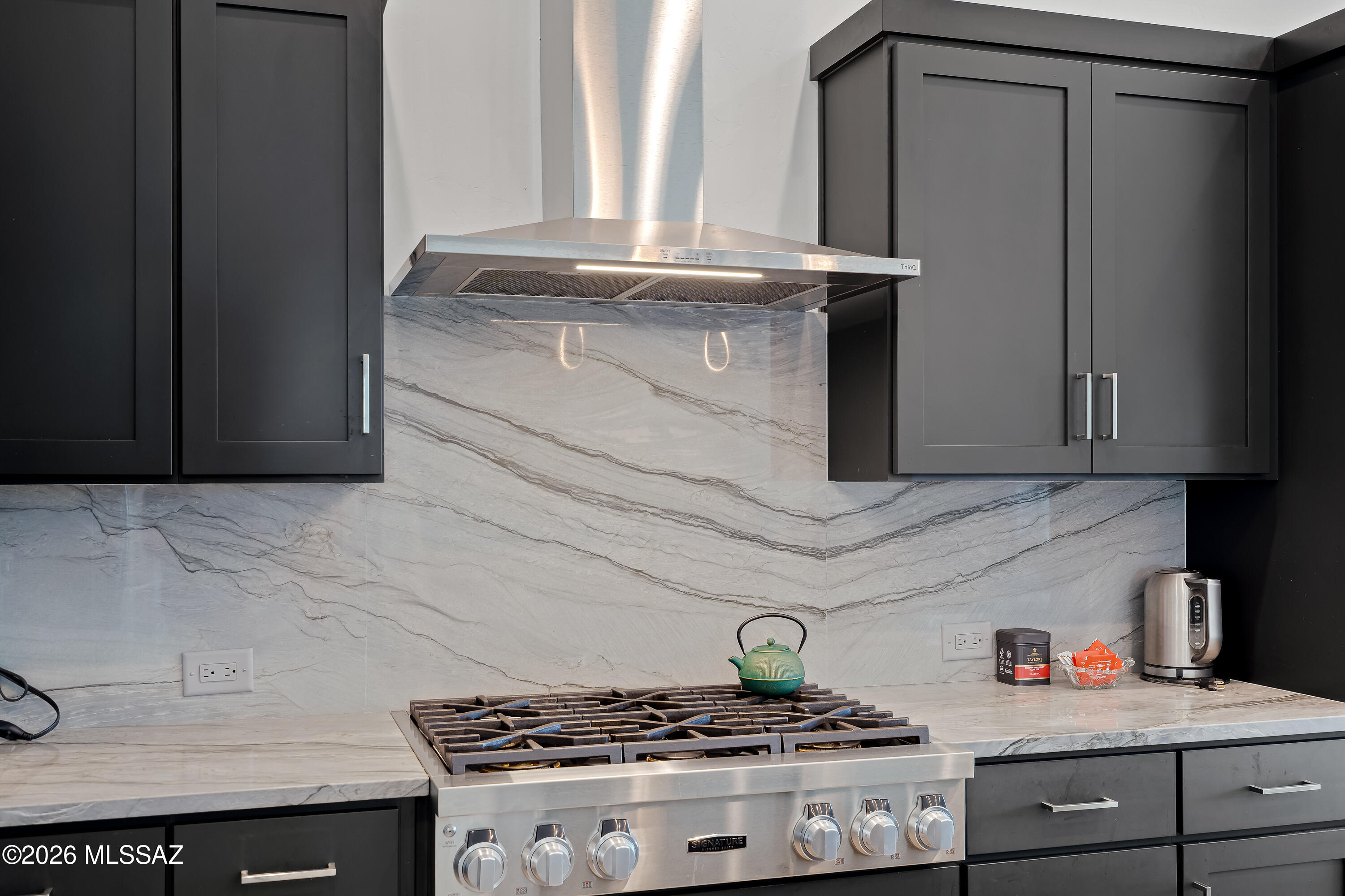Kitchen range beneath a stainless hood, set against dramatic veined stone backsplash and charcoal cabinets.