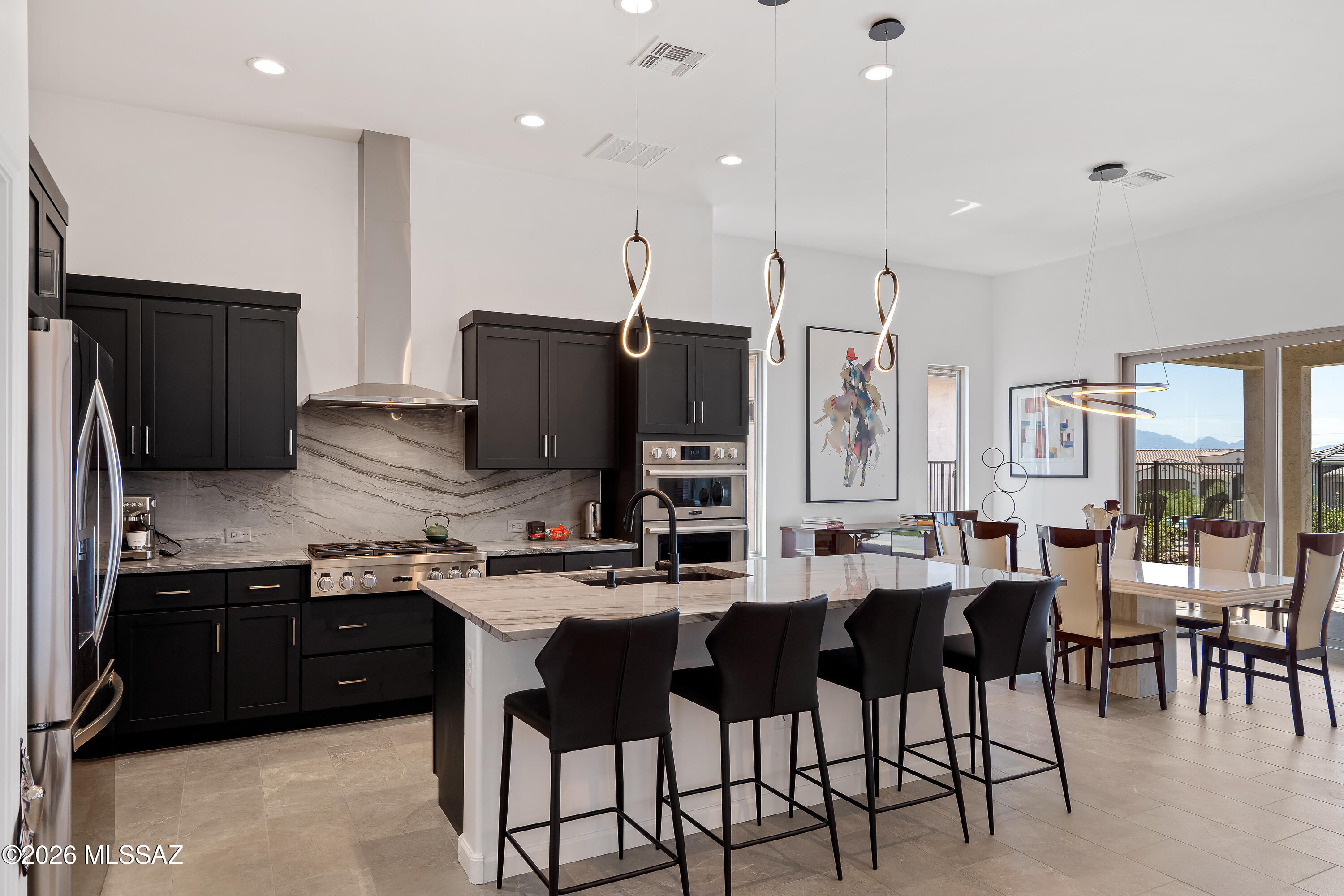 Open kitchen with dark cabinets, stone backsplash, large island, and dining area beside sliding glass doors.