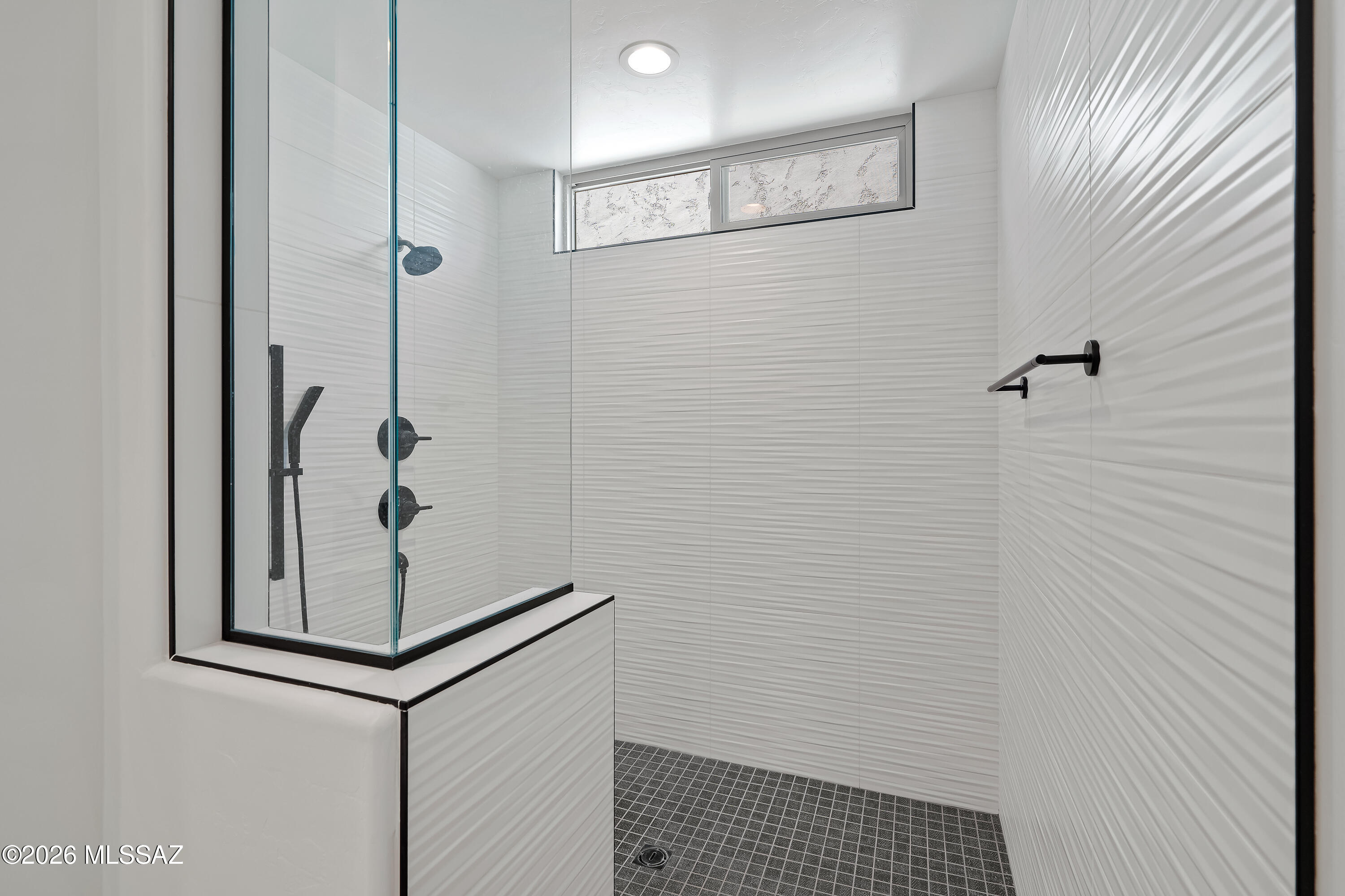 Walk-in shower with white textured tile, glass enclosure, dark fixtures, and dark grid-tile flooring.