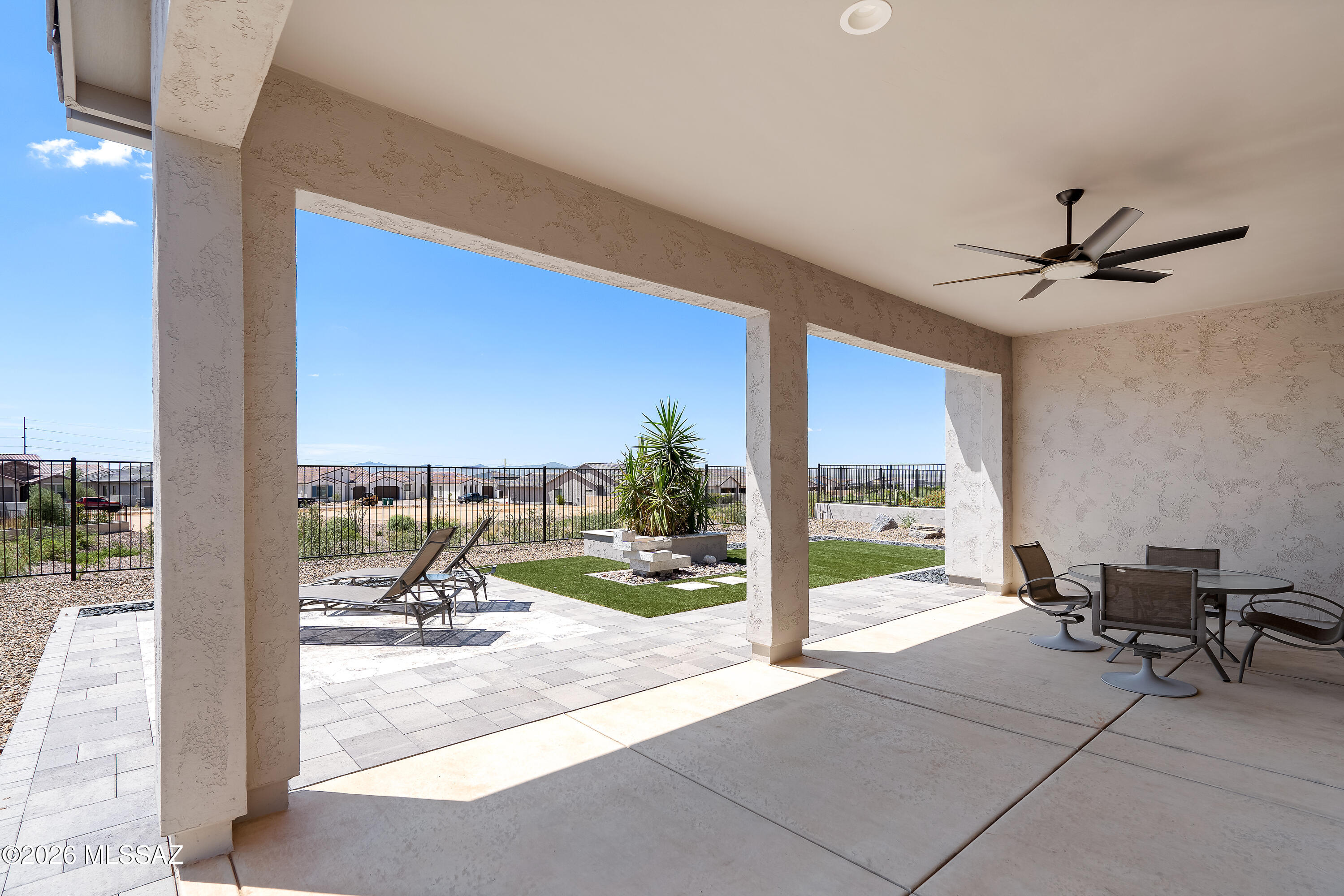 Covered patio with outdoor dining set overlooking a paved backyard with artificial turf and lounge chairs.