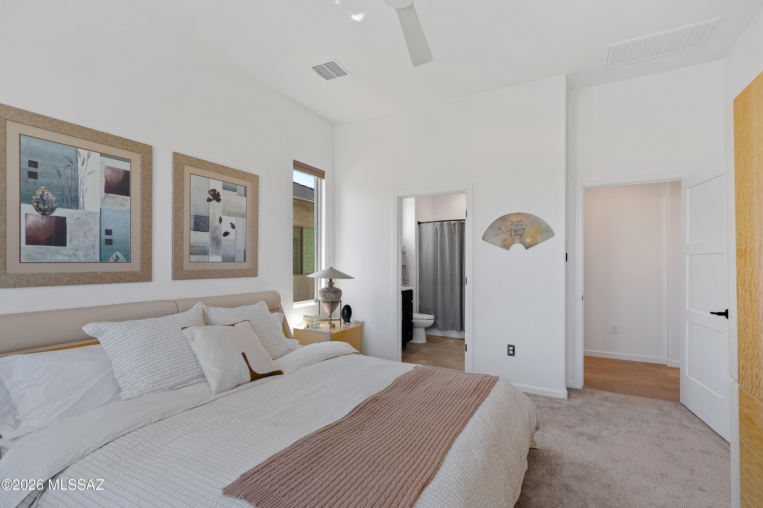 Furnished bedroom with a white bed, dusty rose throw, abstract art, and adjoining bathroom doorway.